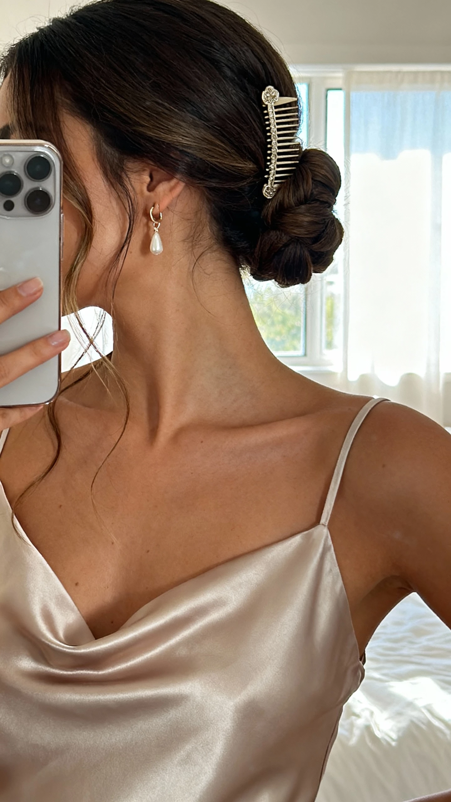 iPhone mirror selfie of a woman in a satin slip dress with delicate drop earrings and a romantic low chignon tucked at the nape, a few soft tendrils and a vintage-inspired hair comb, face not visible, cropped at neck, bedroom mirror with window light, natural iPhone photo quality.