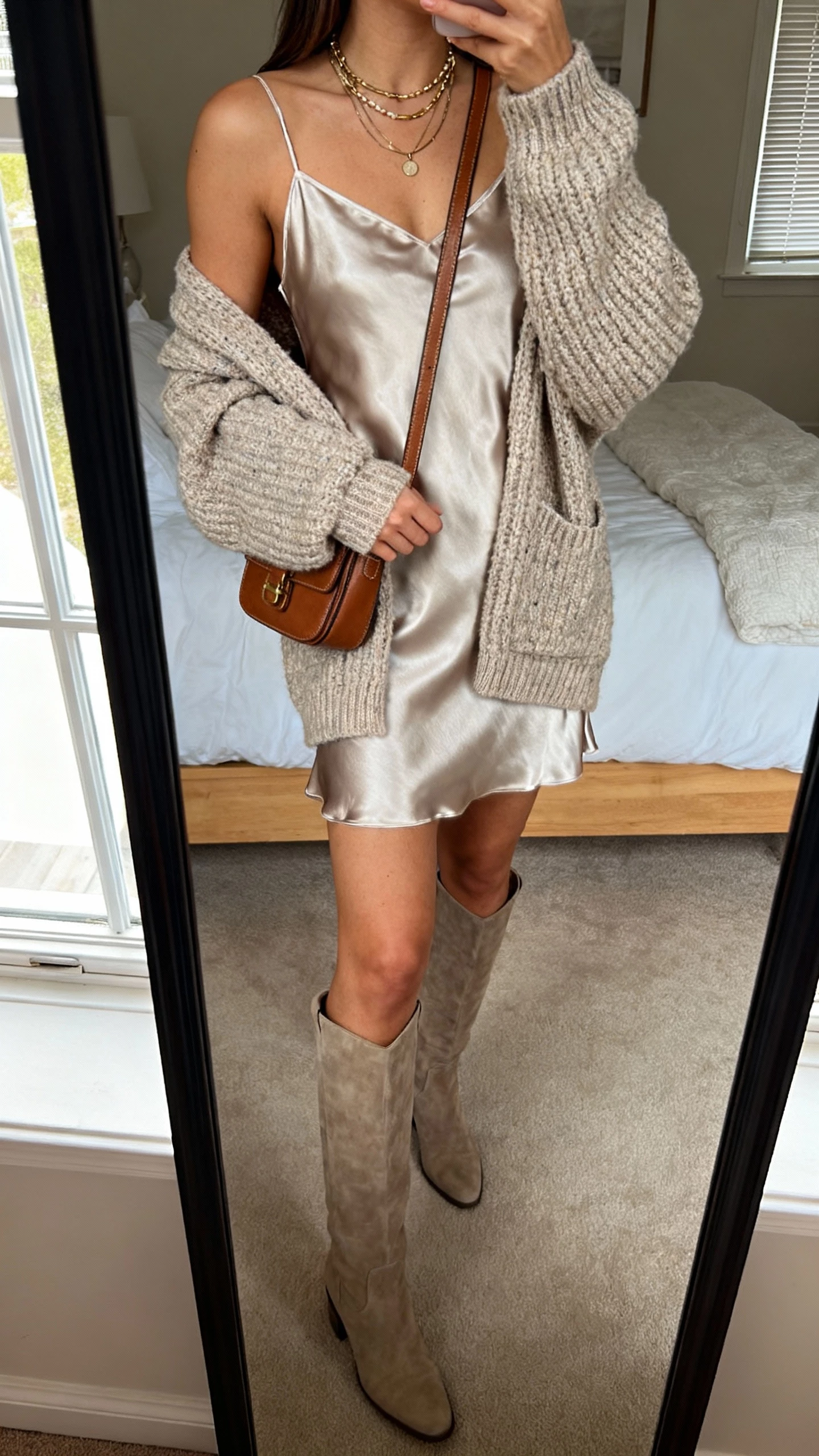 iPhone mirror selfie of a woman in a satin slip dress layered with a chunky cardigan and tall boots, crossbody bag, layered necklaces, face not visible, cropped at shoulders, cozy bedroom mirror, natural window light, iPhone photo quality.