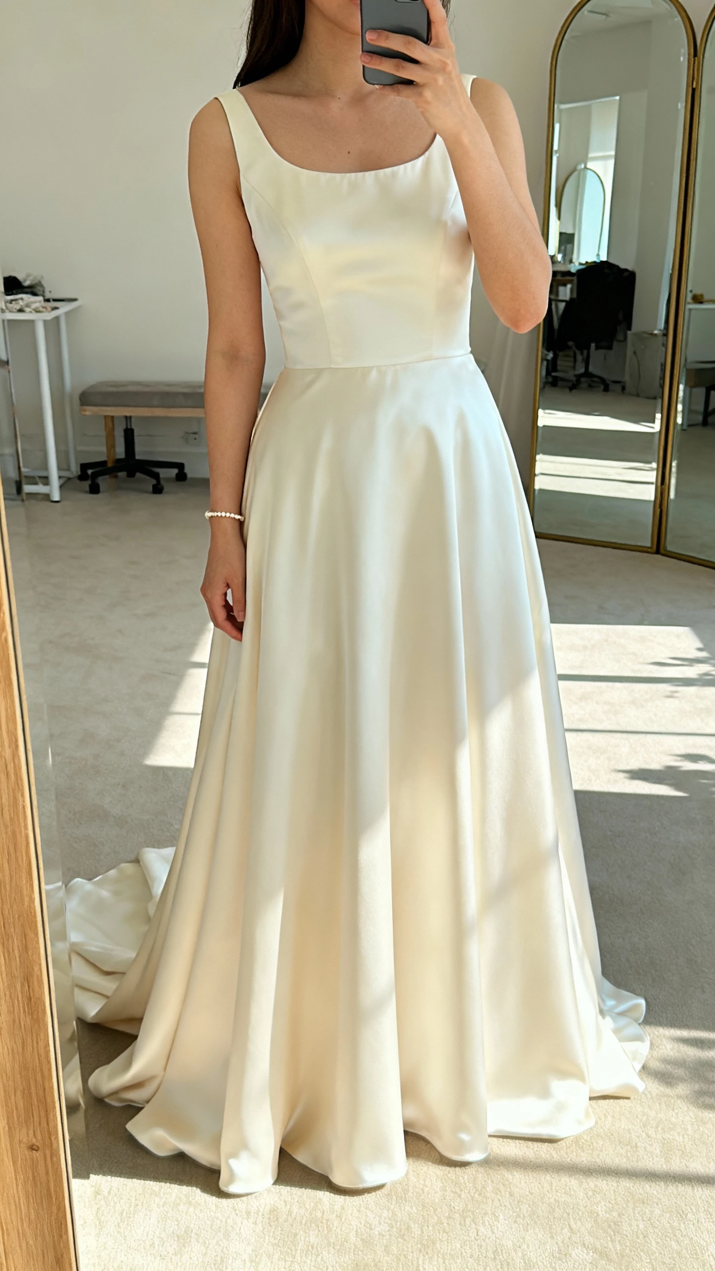iPhone mirror selfie of a woman in a scoop-neck wedding dress with balanced neckline, soft flowy skirt, satin finish, delicate bracelet, face not visible, airy studio mirror, afternoon natural light, casual iPhone photo.