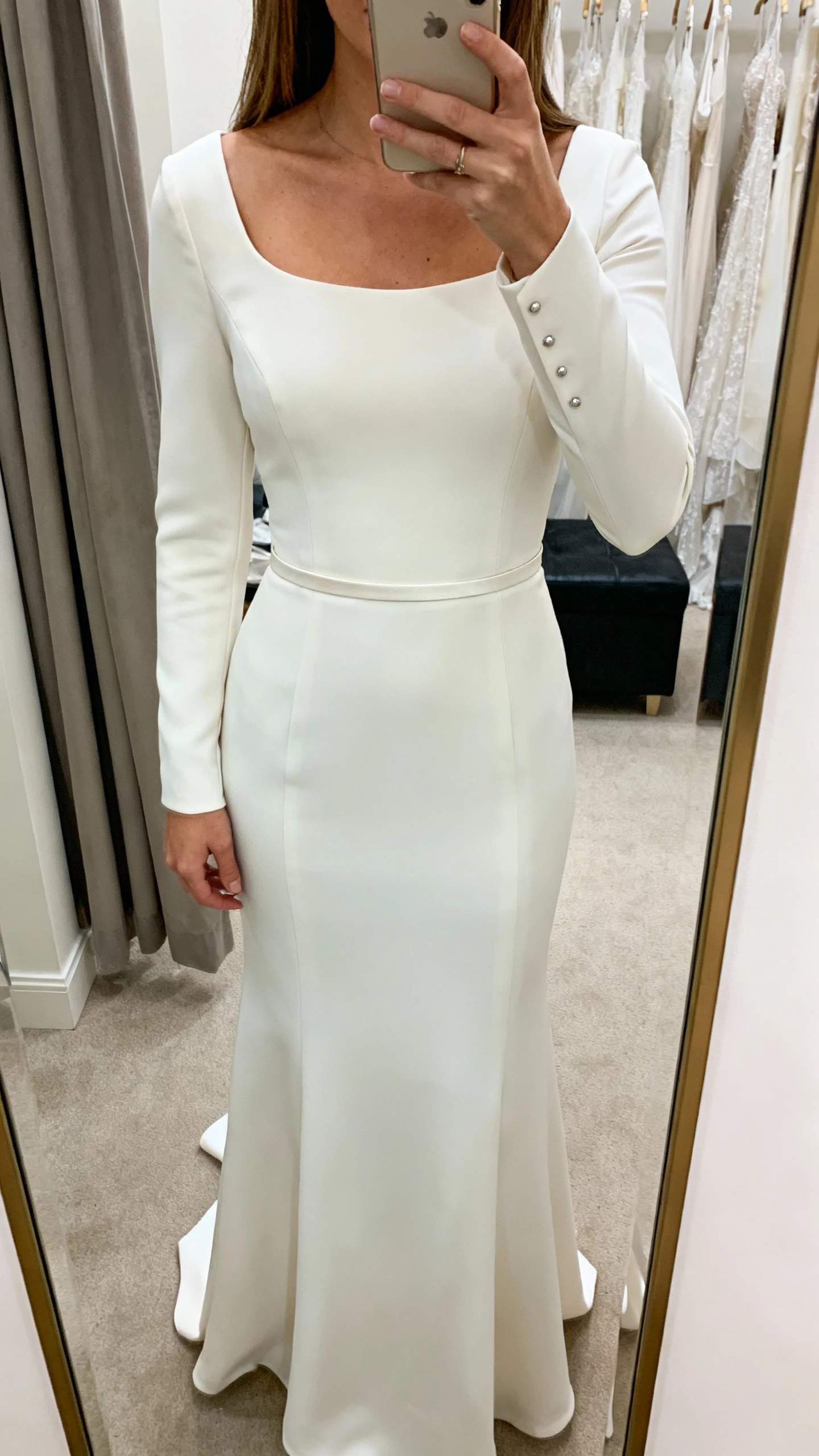iPhone mirror selfie of a woman in a sleek crepe bridal gown with clean lines, fitted bodice, subtle scoop neckline, long sleeves, minimalist studs, thin crepe belt, face not visible, cropped at shoulders, dressing room mirror, natural indoor light, iPhone photo quality