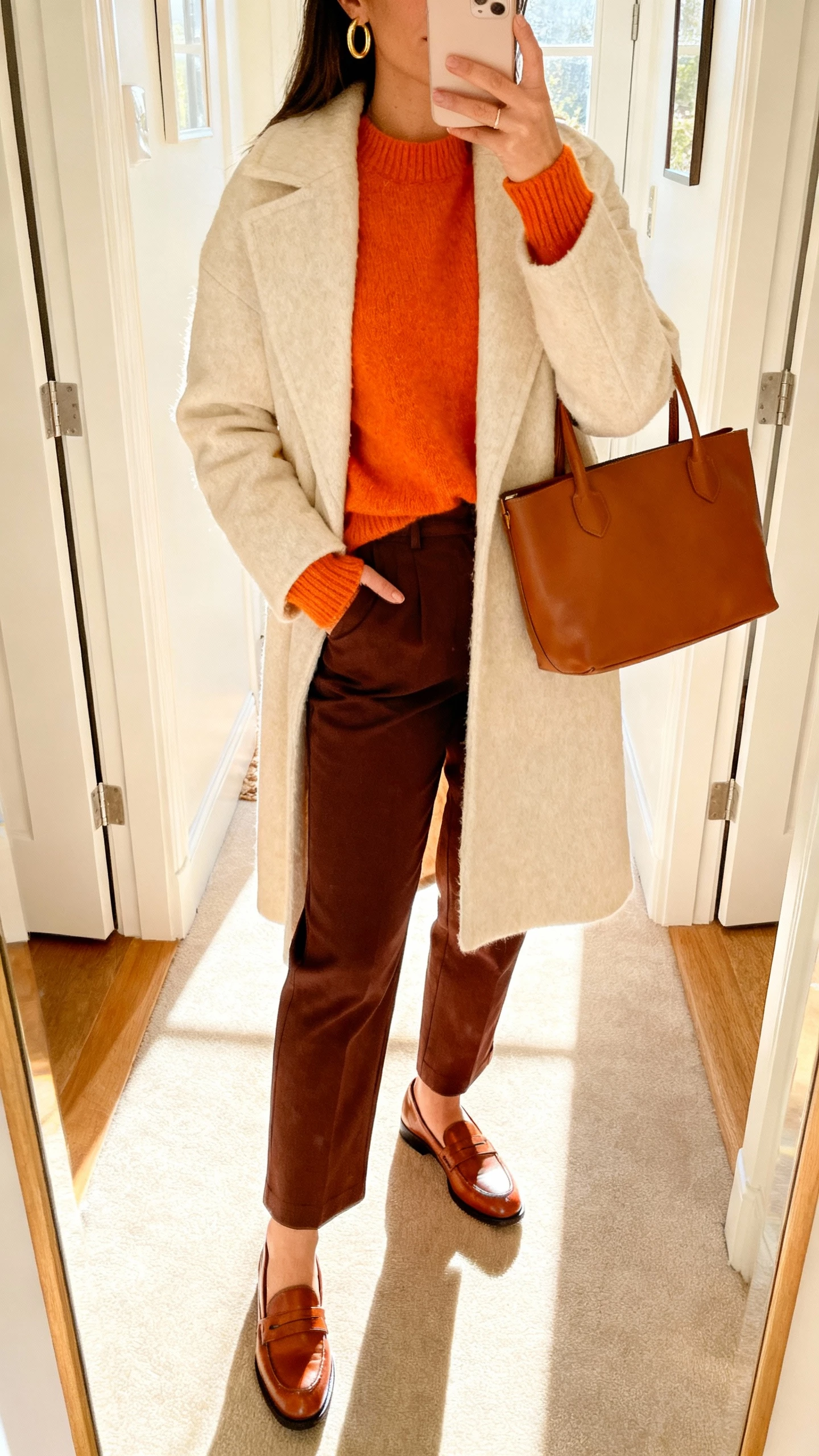 iPhone mirror selfie of a woman in a tangerine sweater, cream wool coat, chocolate brown trousers, brown leather loafers and tote, subtle gold hoops, face not visible, entryway mirror with bright morning light, natural iPhone aesthetic