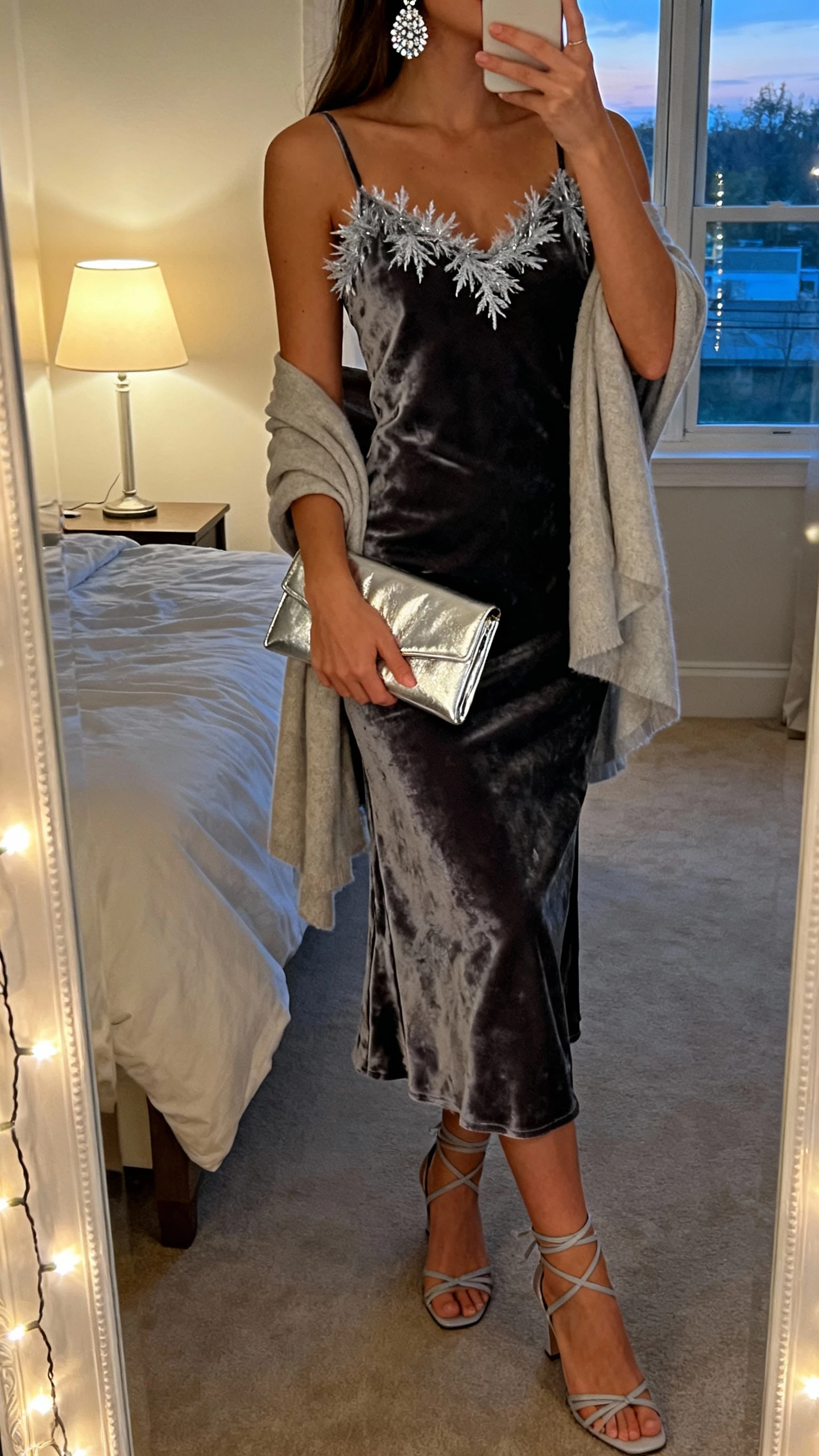 iPhone mirror selfie of a woman in a velvet slip dress with frosty silver accents—sparkly earrings, metallic clutch, strappy heels, and a soft shawl—face not visible, cropped at neck, bedroom mirror with fairy lights, natural evening lamp + window light, natural iPhone photo quality.