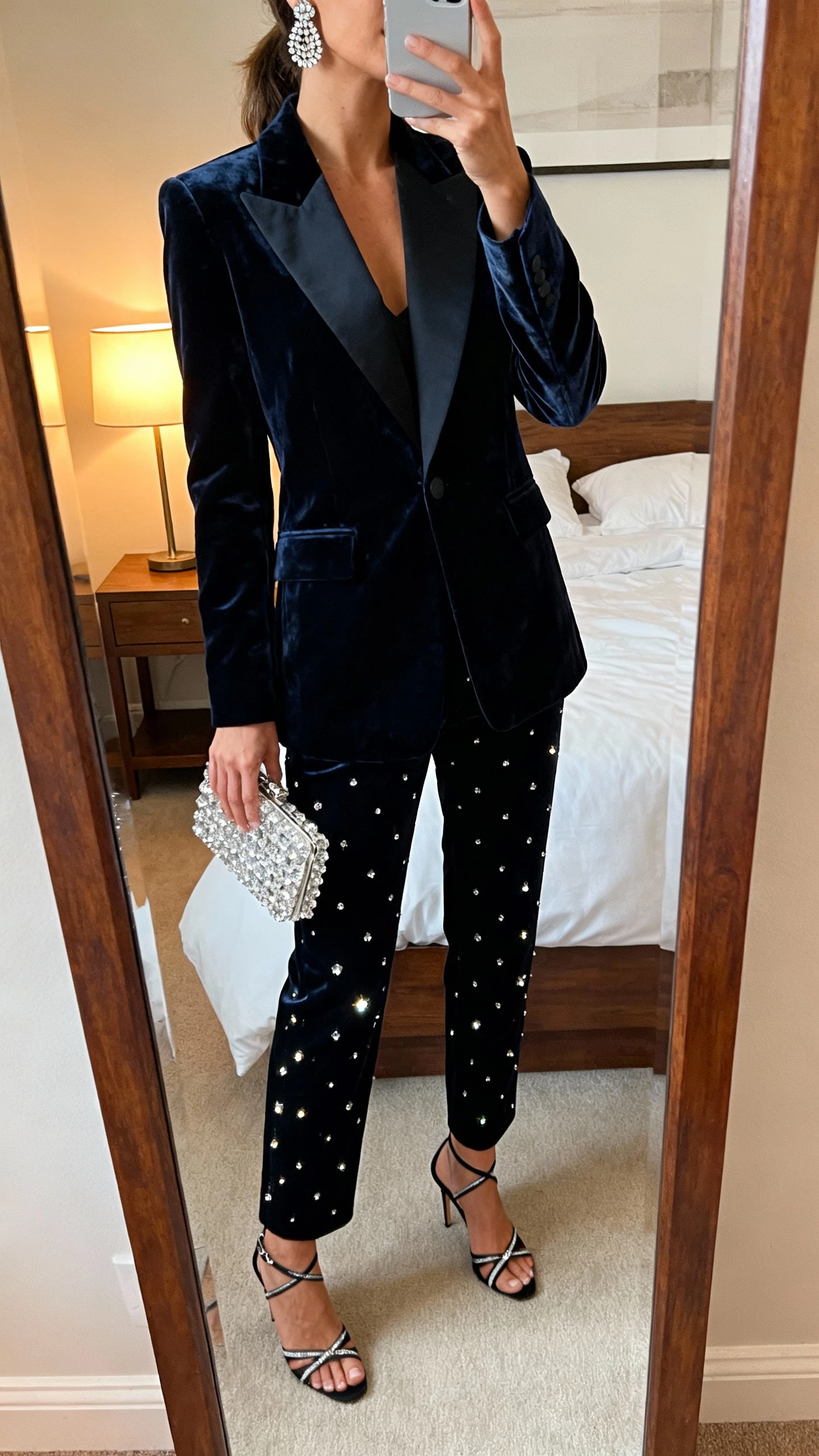 iPhone mirror selfie of a woman in a velvet suit (tailored blazer and trousers) with sparkly accents, strappy heels, rhinestone earrings, and a crystal clutch, face not visible, cropped at neck, bedroom mirror with warm lamp light, natural iPhone photo quality.