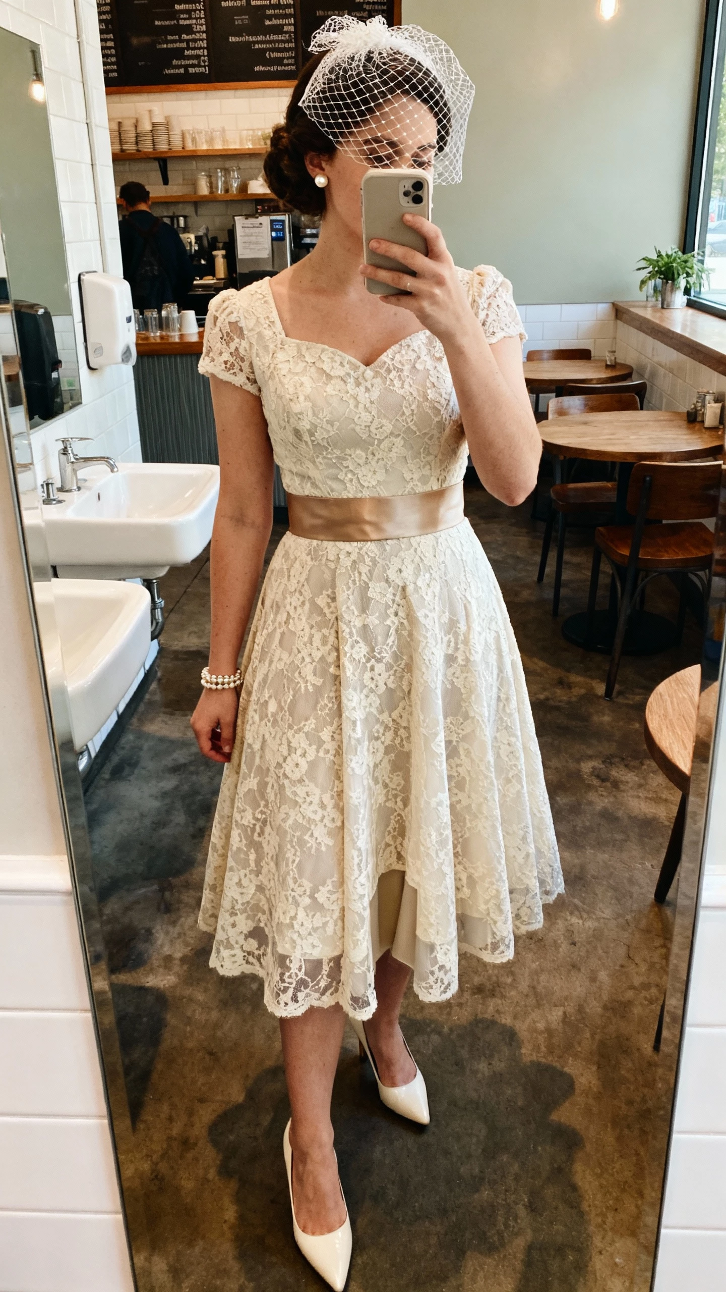 iPhone mirror selfie of a woman in a vintage-inspired tea-length lace dress with a structured bodice and A-line skirt, wide satin belt, birdcage veil, pointed kitten heels, pearl studs, vintage-style bracelet, lightweight slip with smoothing panel; chic cafe restroom mirror, face not visible, warm indoor lighting, natural iPhone photo quality.