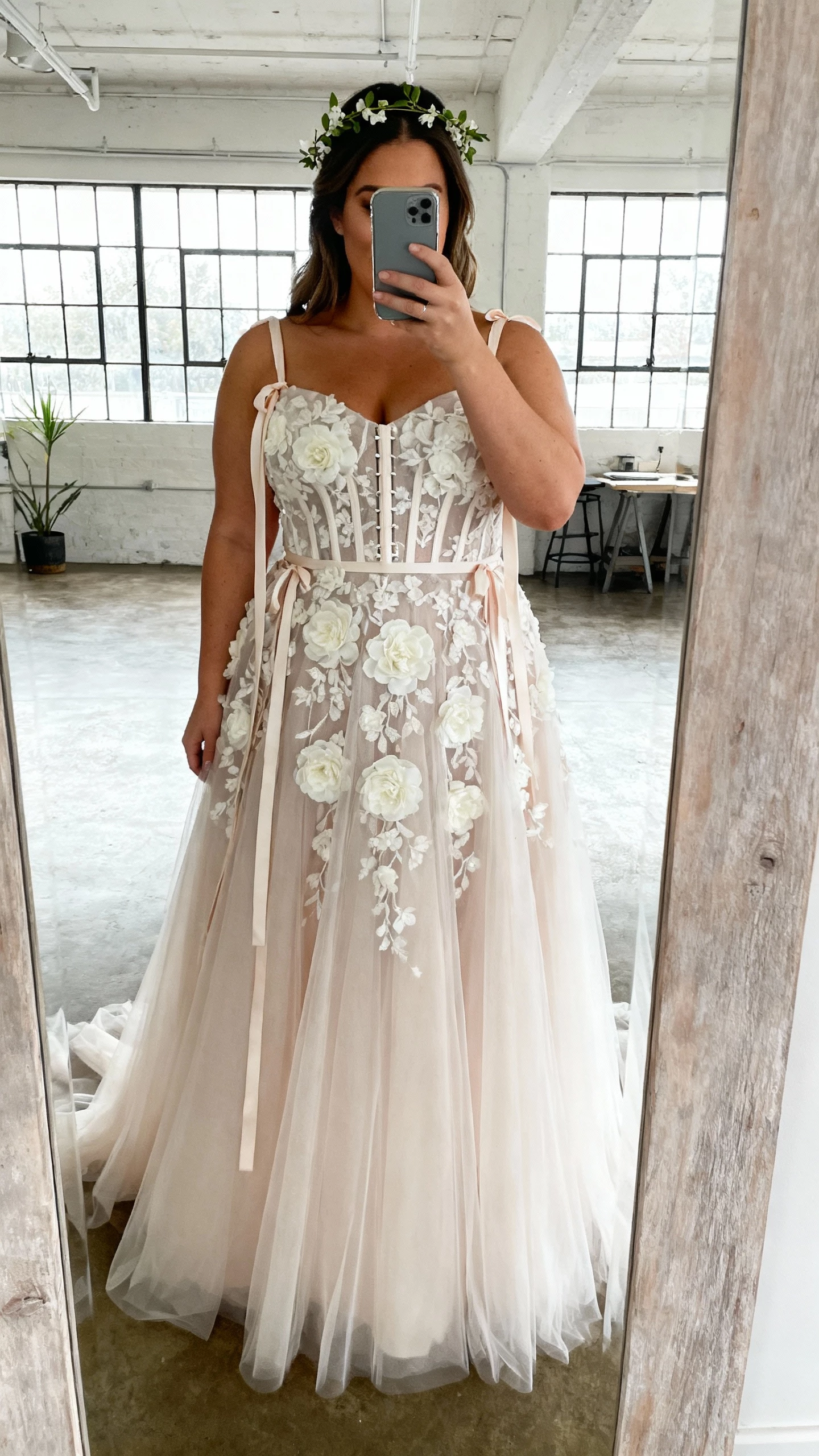 iPhone mirror selfie of a woman in a Watters Wtoo Curve airy tulle A-line with dimensional floral appliqués, corset-style bodice, and soft ribbon straps, paired with delicate vine headband; face not visible, studio loft mirror, diffused window light, casual iPhone aesthetic.