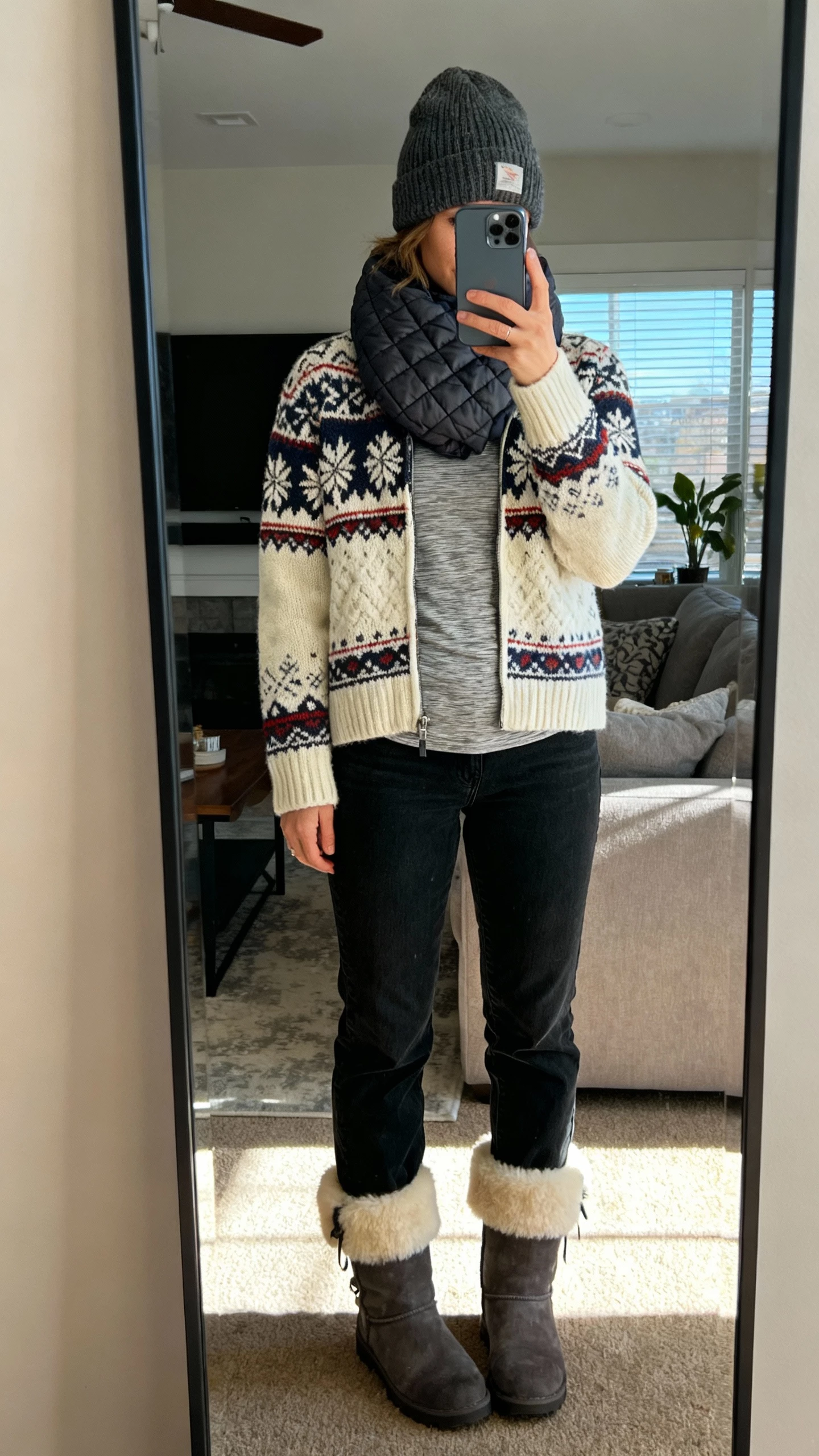 iPhone mirror selfie of a woman in alpine-in-the-city knitwear: Fair Isle sweater over a thermal tee, wool beanie, straight black jeans, shearling-lined boots, and a quilted scarf, face not visible, cozy living room mirror with afternoon light, casual iPhone quality.