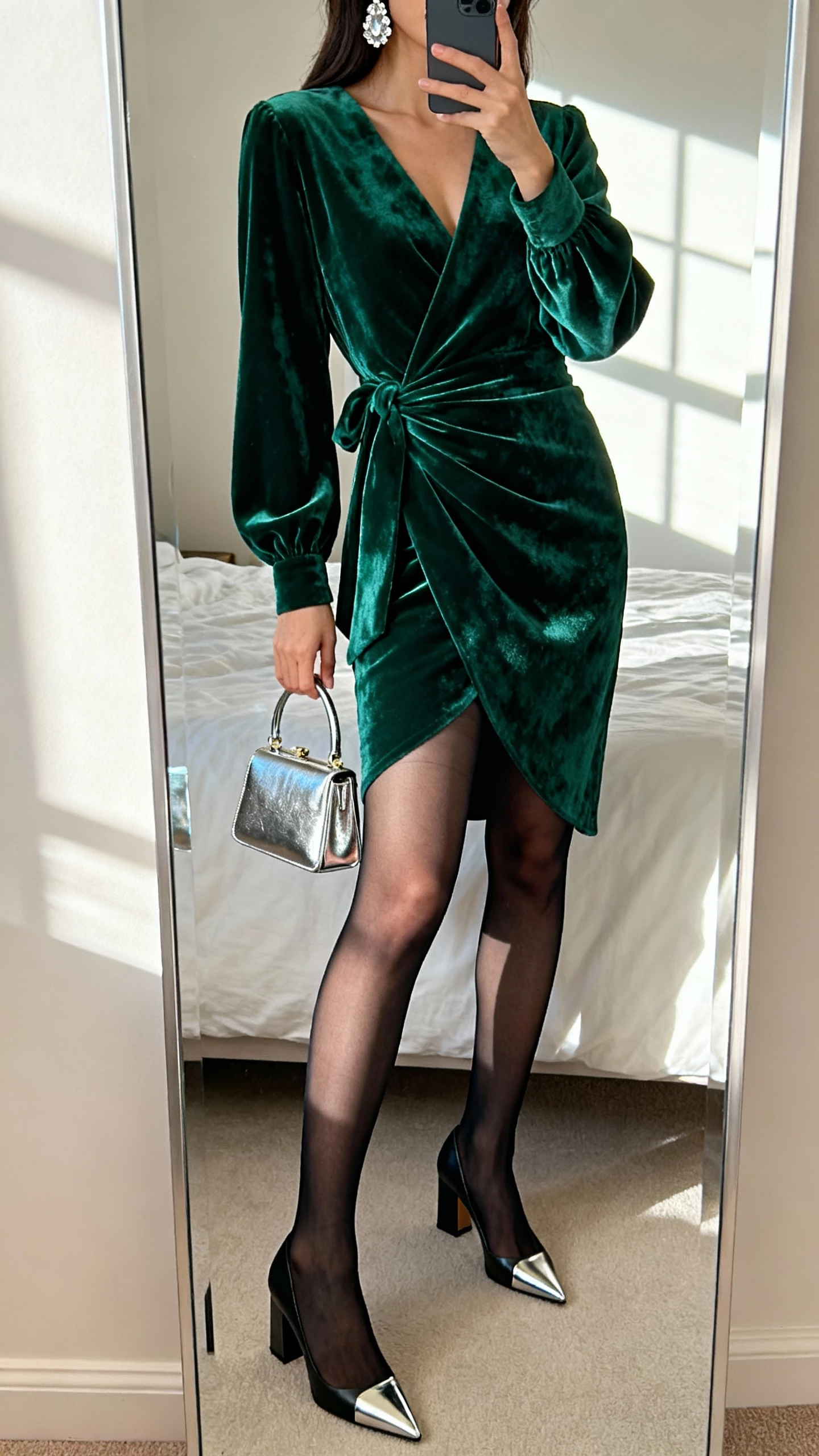 iPhone mirror selfie of a woman in an emerald velvet wrap dress with long sleeves, sheer black tights, pointed-toe block-heel pumps, crystal drop earrings, and a metallic mini top-handle bag; face not visible, cropped at neck, bedroom mirror with window light, natural iPhone photo quality.