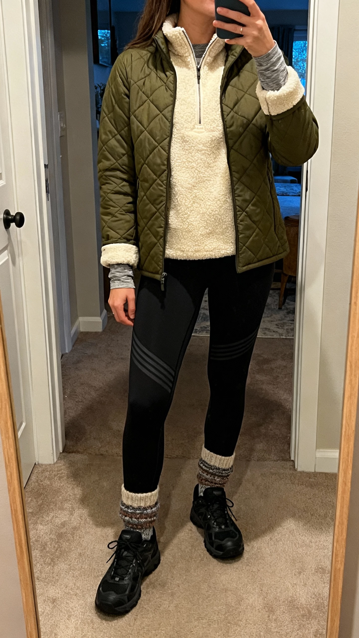 iPhone mirror selfie of a woman in an olive quilted liner jacket over a high-pile cream fleece half-zip, thermal-lined black leggings, water-resistant black sneakers, thick wool socks with subtle pattern, base-layer long-sleeve visible at cuffs, face not visible, cozy entryway mirror with indoor evening light, natural iPhone photo quality.