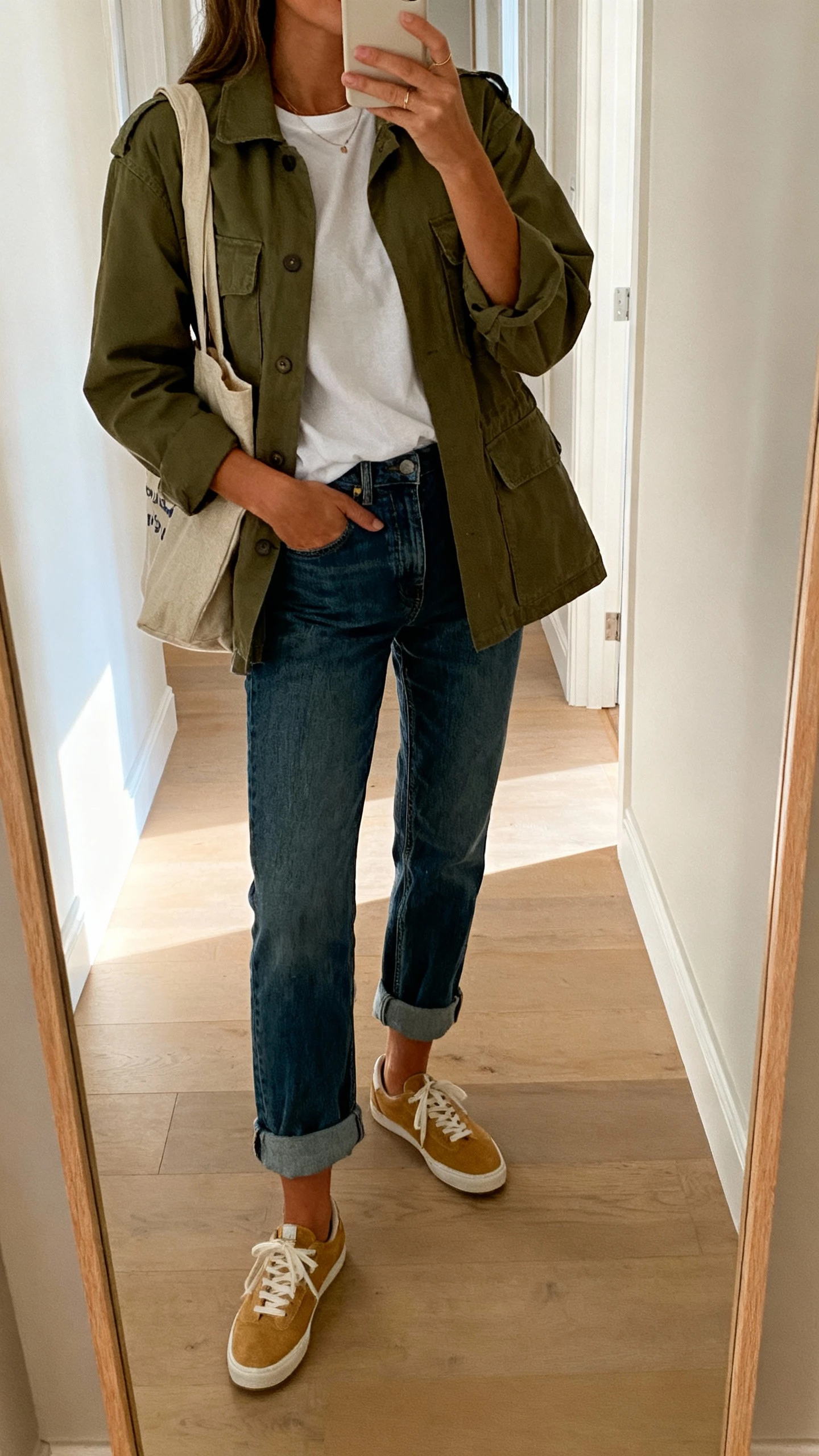 iPhone mirror selfie of a woman in an olive utility jacket over a white tee with boyfriend jeans, rolled cuffs, tan sneakers, canvas tote, face not visible, hallway mirror, natural afternoon light, iPhone photo quality.