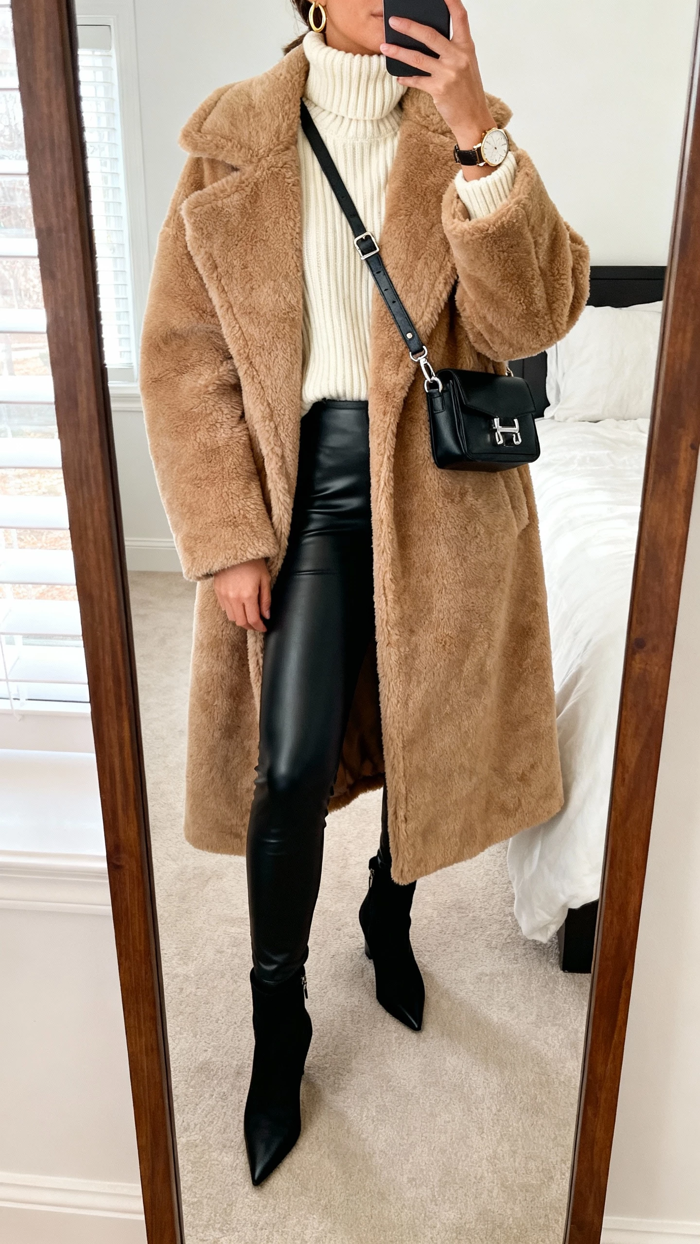 iPhone mirror selfie of a woman in an oversized camel teddy coat, black faux‑leather leggings, chunky cream ribbed turtleneck, pointed ankle boots, structured crossbody with metal hardware, minimal gold hoops and a statement watch; bedroom full-length mirror, face not visible, natural window light, natural iPhone photo quality.