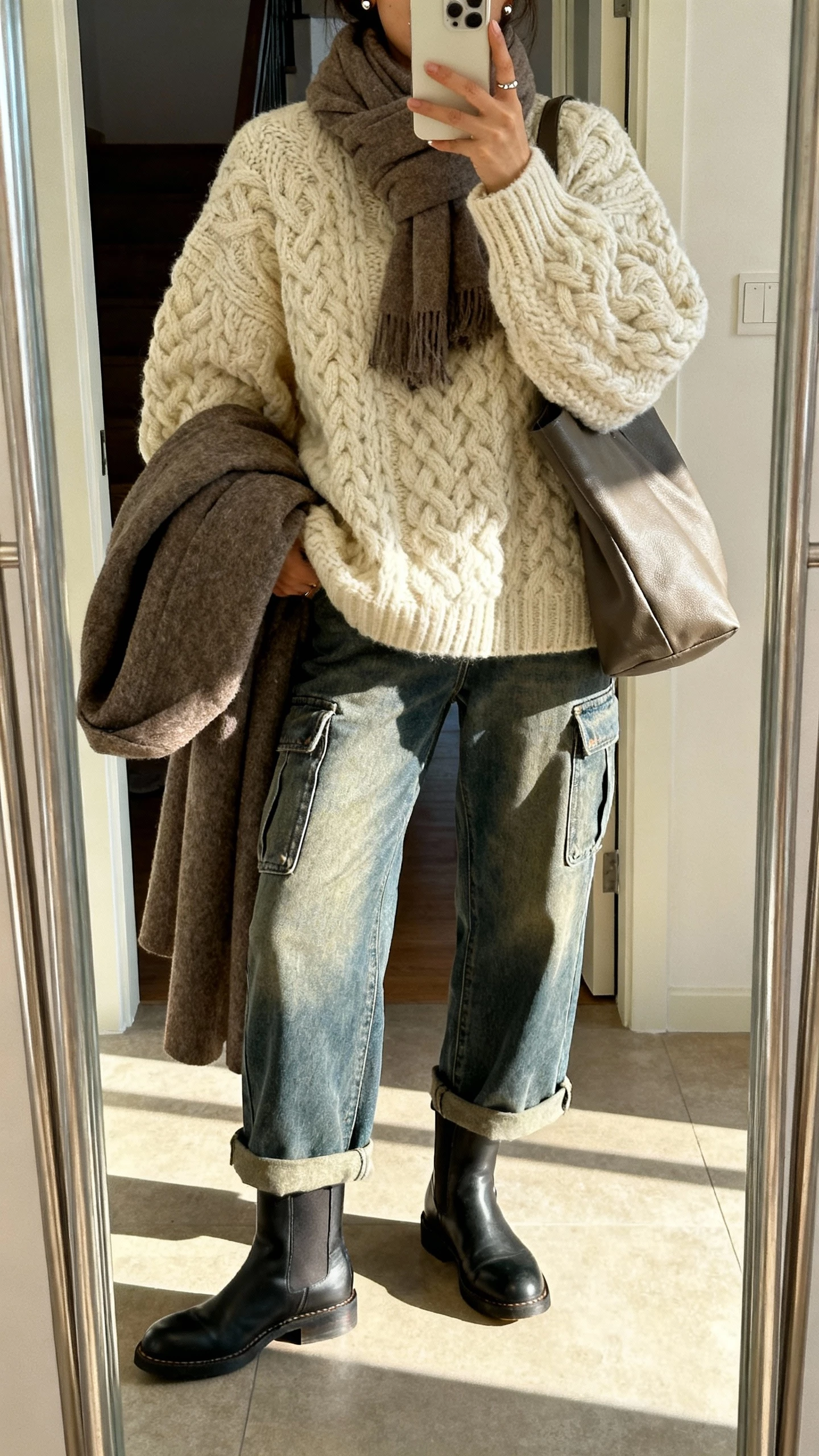 iPhone mirror selfie of a woman in an oversized chunky knit sweater in cream, relaxed mid-length cargo jeans, Chelsea boots, wool coat draped over arm, cozy scarf, minimal studs, and a slouchy tote, face not visible, entryway mirror, afternoon natural light, iPhone photo quality.