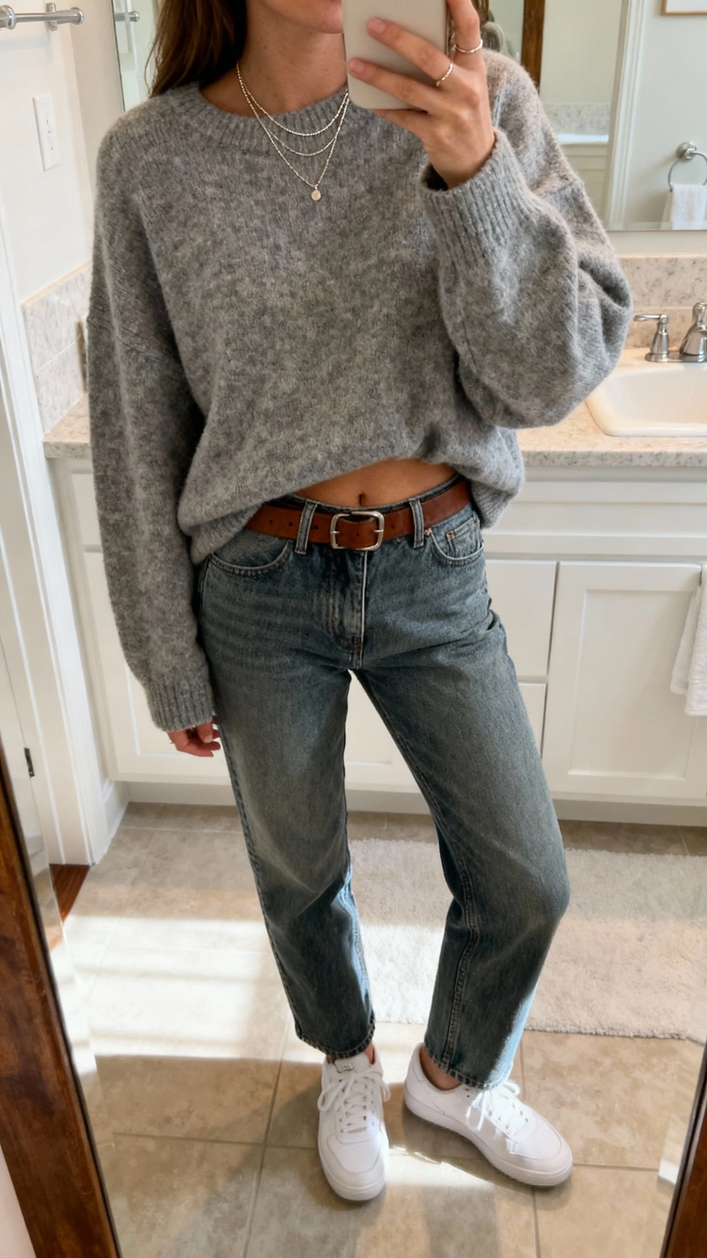 iPhone mirror selfie of a woman in an oversized heather-gray sweater half-tucked into straight-leg medium-wash jeans, white sneakers, leather belt, layered dainty necklaces, face not visible, bathroom mirror, soft morning light, natural iPhone photo quality.