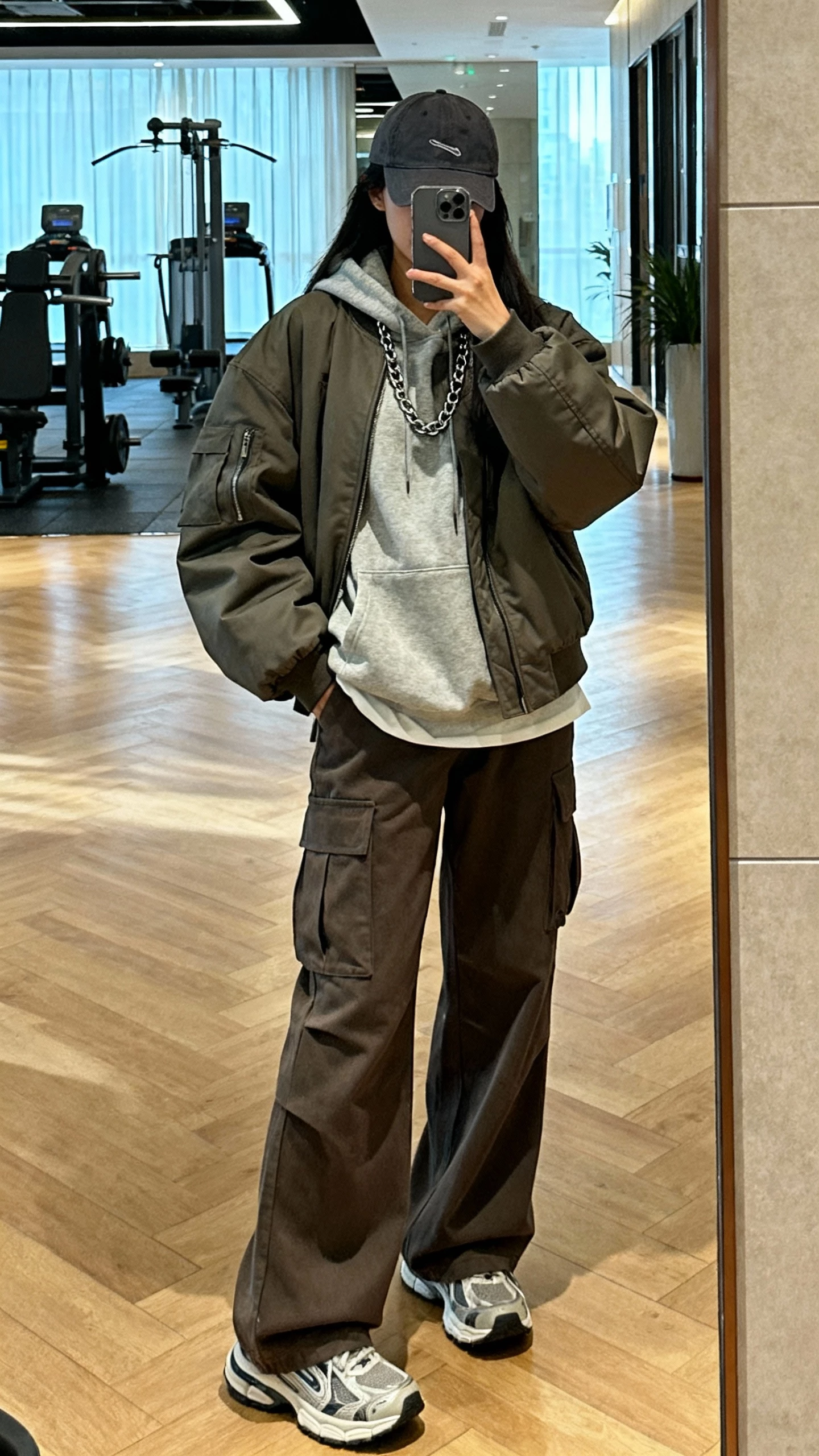 iPhone mirror selfie of a woman in elevated streetwear layers: oversized hoodie under a boxy bomber, wide-leg cargo trousers, dad sneakers, baseball cap, and layered chain, face not visible, full-length gym lobby mirror with morning light, casual iPhone look.