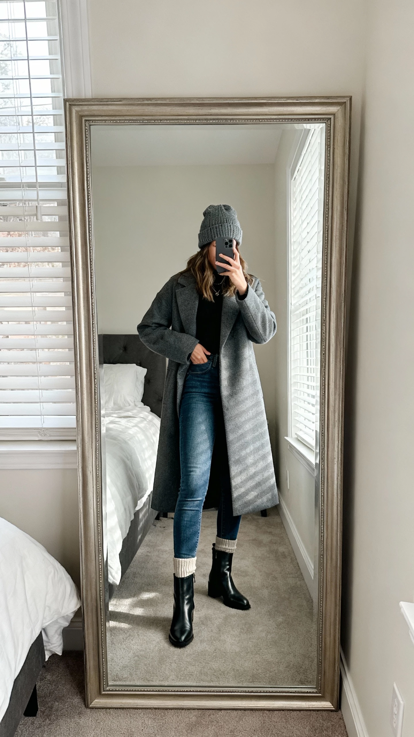 iPhone mirror selfie of a woman in fitted jeans tucked into sleek ankle-covering boots, ribbed socks hidden, long coat, knit beanie, face not visible, full-length mirror in bedroom, natural window light, iPhone photo quality.