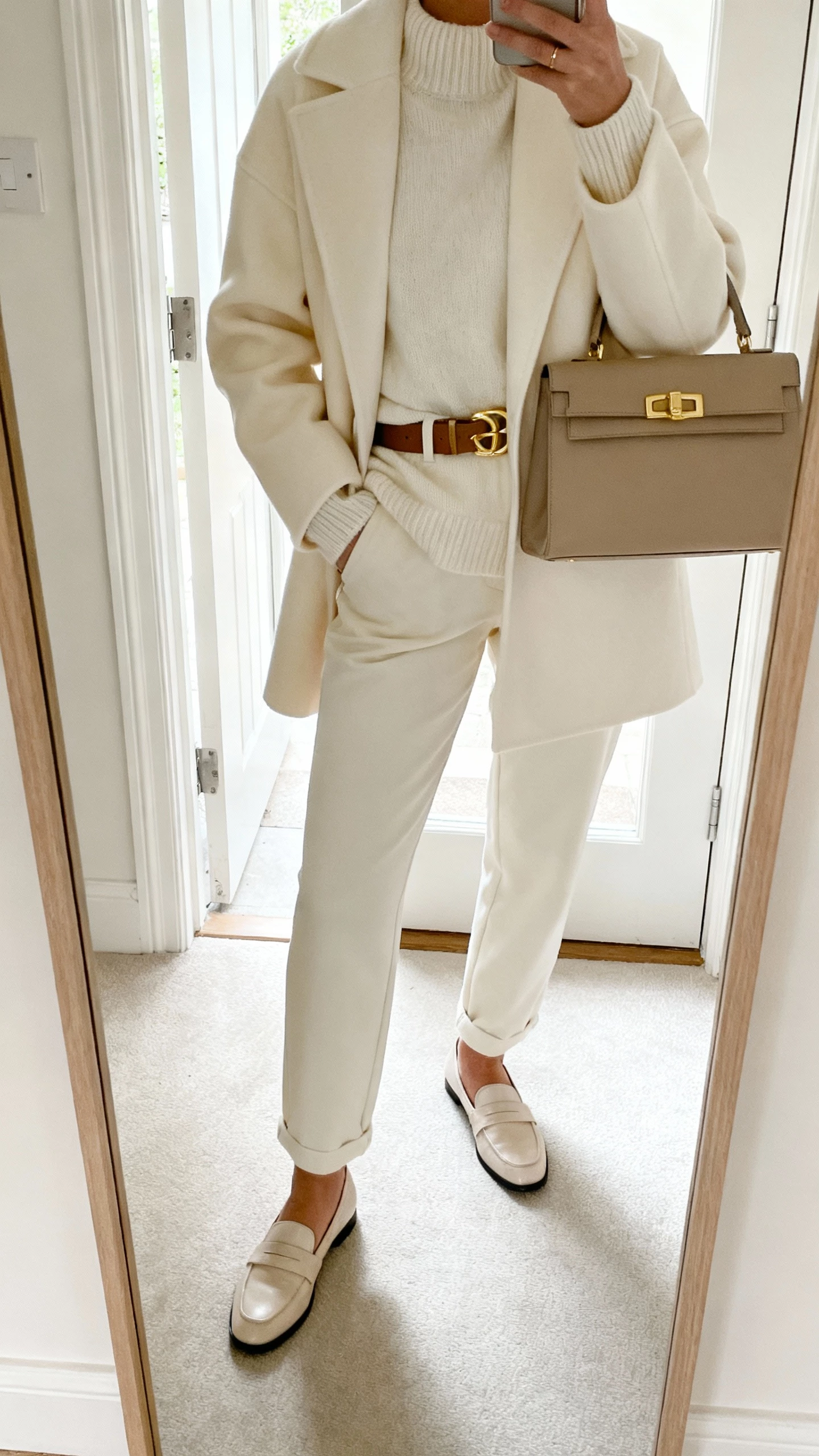 iPhone mirror selfie of a woman in monochrome cream layers (coat, knit, trousers) with gold hardware on belt and bag, plus loafers, face not visible, entryway mirror with daylight from door, natural iPhone photo quality.