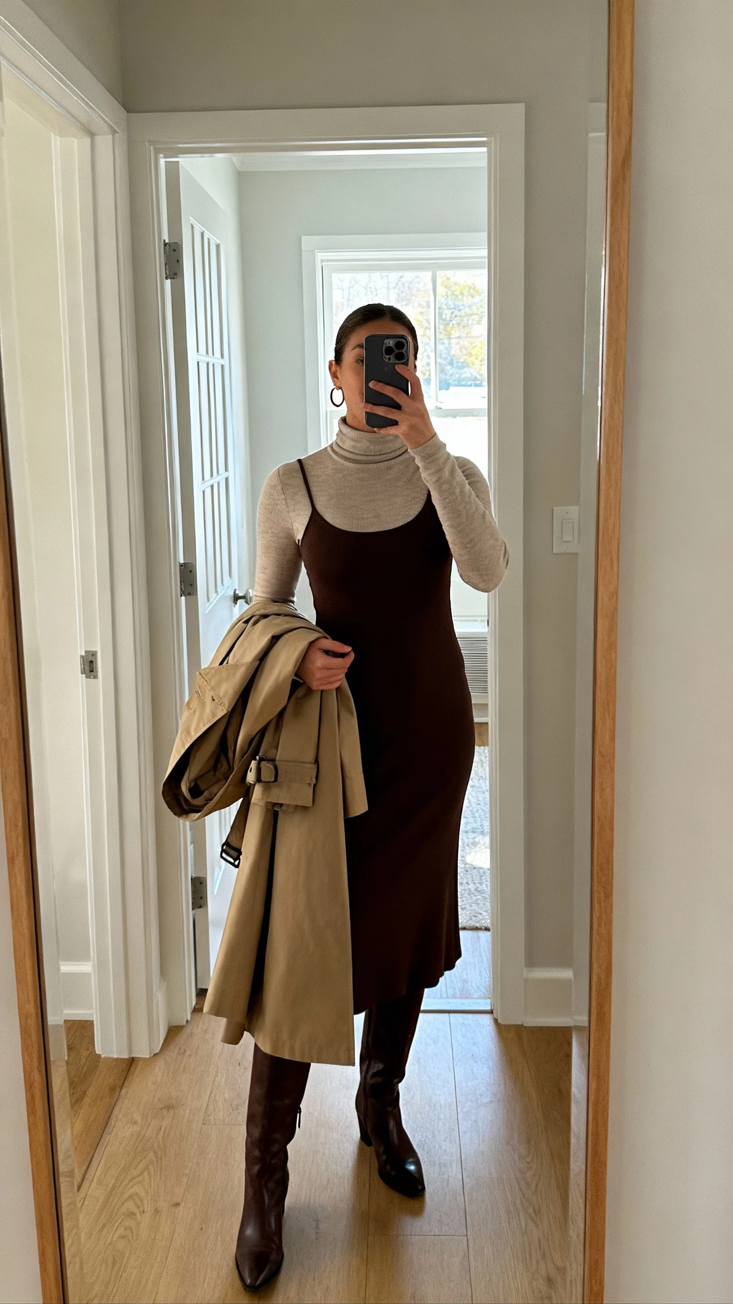 iPhone mirror selfie of a woman layering a fitted turtleneck under a midi dress with knee-high boots, trench draped over arm, simple hoops, face not visible, entryway mirror, morning window light, natural iPhone aesthetic.