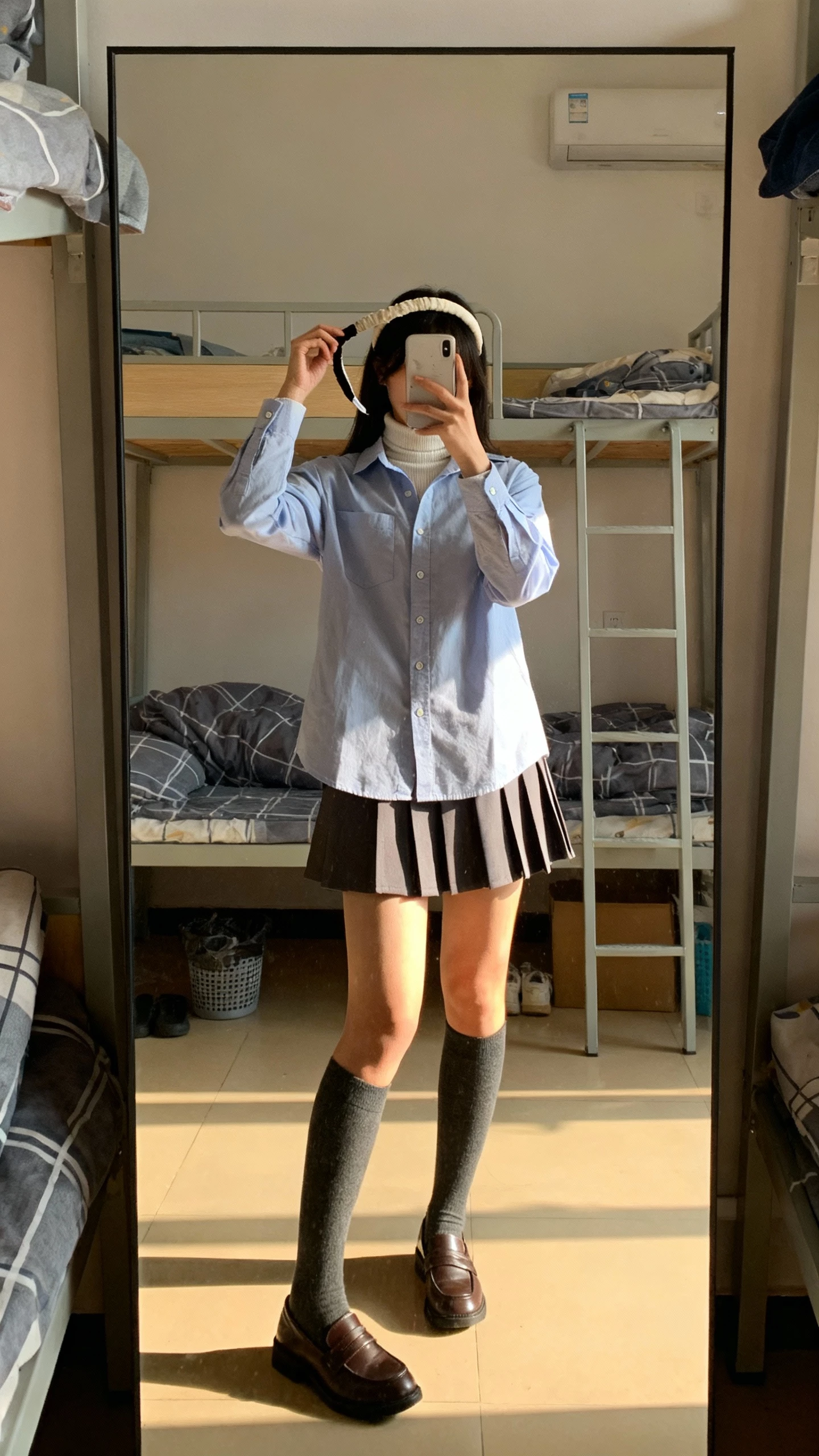iPhone mirror selfie of a woman layering a turtleneck under an Oxford button-down with a pleated mini skirt, knee socks and loafers, headband in hand, face not visible, dorm-style bedroom mirror, afternoon light, natural iPhone photo quality.