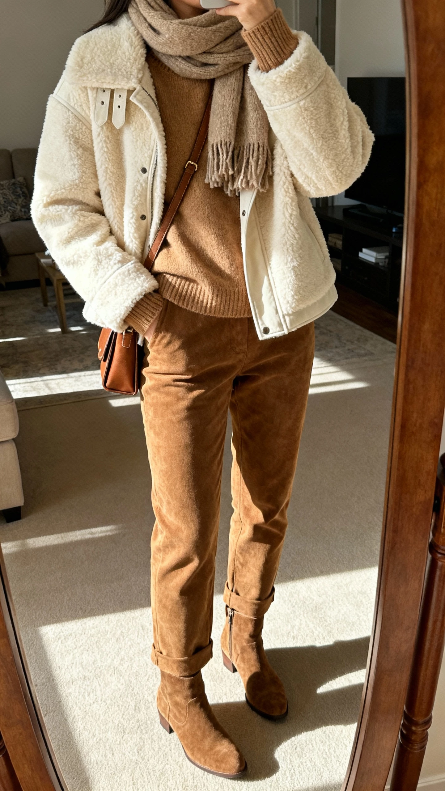 iPhone mirror selfie of a woman layering tonal textures: cream shearling jacket over a cashmere camel sweater, tan suede trousers, suede ankle boots, wool scarf, leather crossbody, face not visible, cropped at shoulders, living room mirror with morning light, natural iPhone aesthetic.