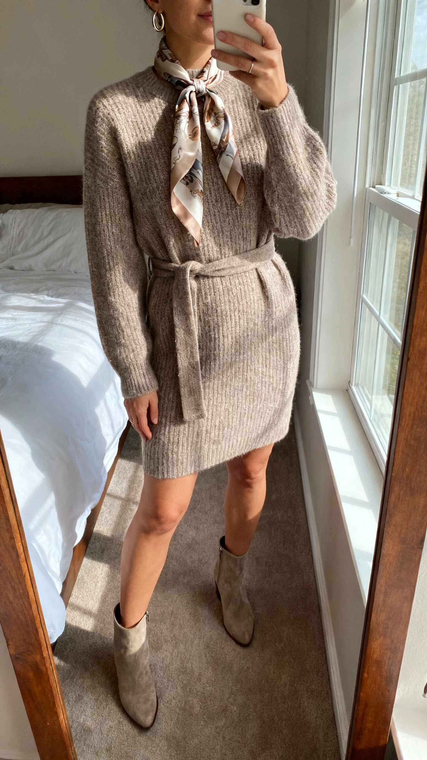 iPhone mirror selfie of a woman wearing a belted sweaterdress with a silky printed scarf tied at the neck, ankle boots, simple hoop earrings, face not visible, cropped at neck, bedroom mirror with morning window light, natural iPhone photo quality.