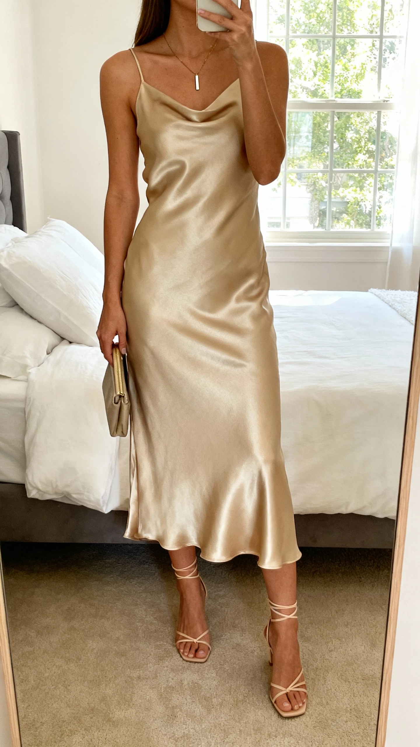 iPhone mirror selfie of a woman wearing a bias-cut slip midi dress in champagne-tone satin with delicate strappy heels, slim pendant necklace, and a small clutch, face not visible, cropped at neck, bedroom mirror with morning window light, natural iPhone photo quality.
