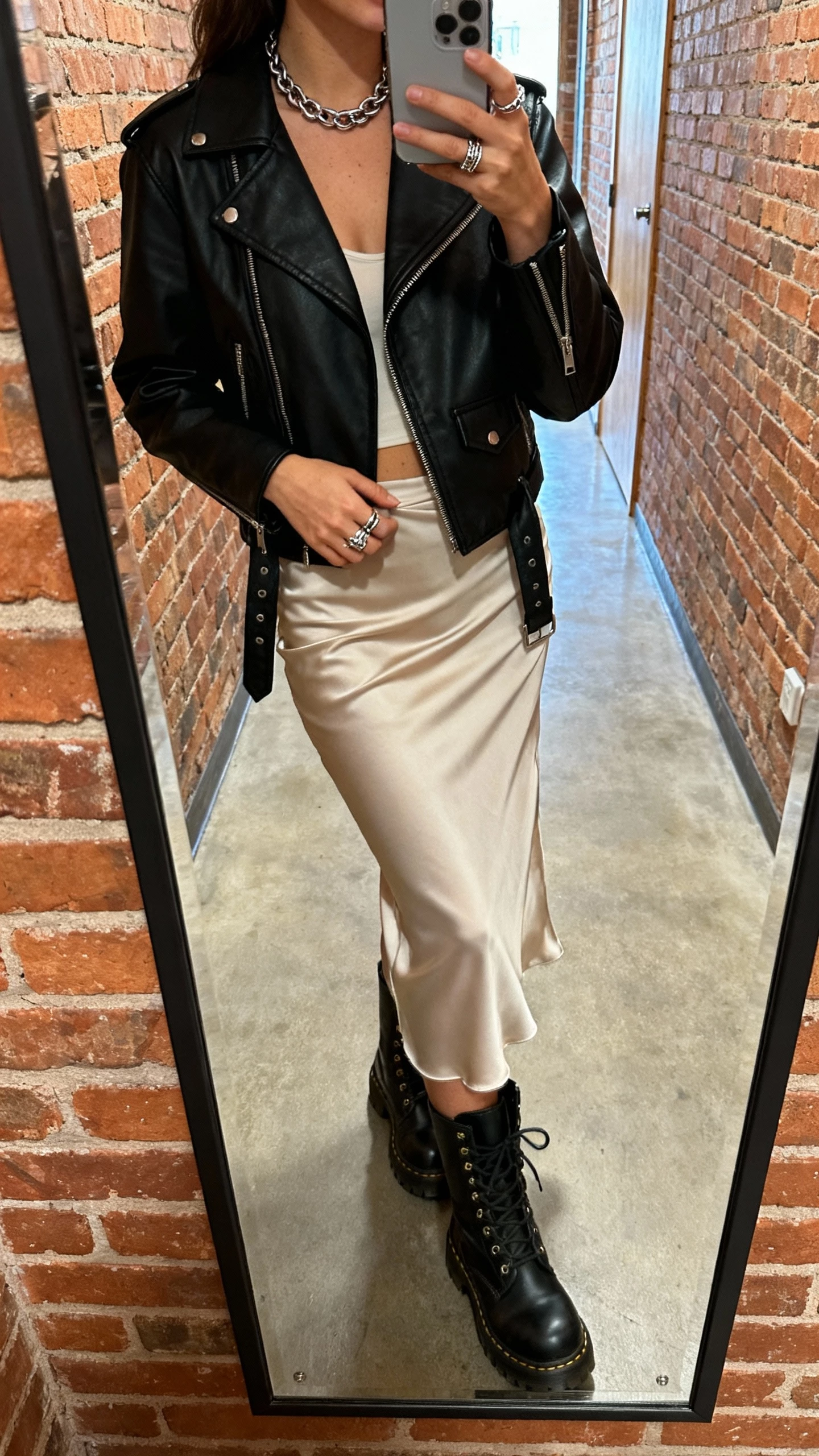 iPhone mirror selfie of a woman wearing a bias-cut slip skirt with chunky combat boots and a black moto jacket, chain necklace and ring set, face not visible, brick-wall hallway mirror, natural iPhone photo quality