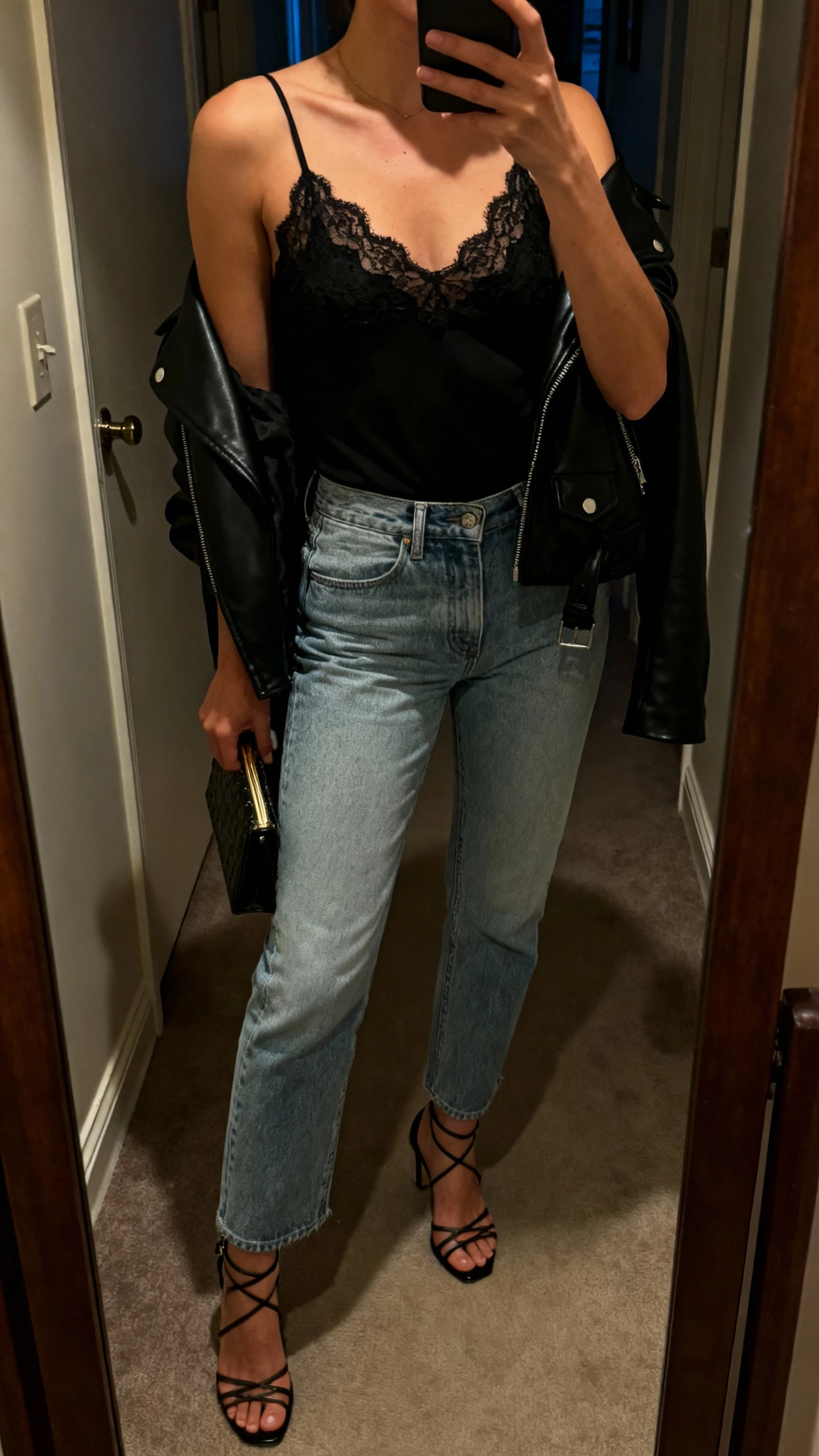 iPhone mirror selfie of a woman wearing a black lace camisole with light blue jeans, black leather moto jacket draped over shoulders, strappy heels, small clutch, face not visible, dim hallway mirror with warm evening light, natural iPhone aesthetic