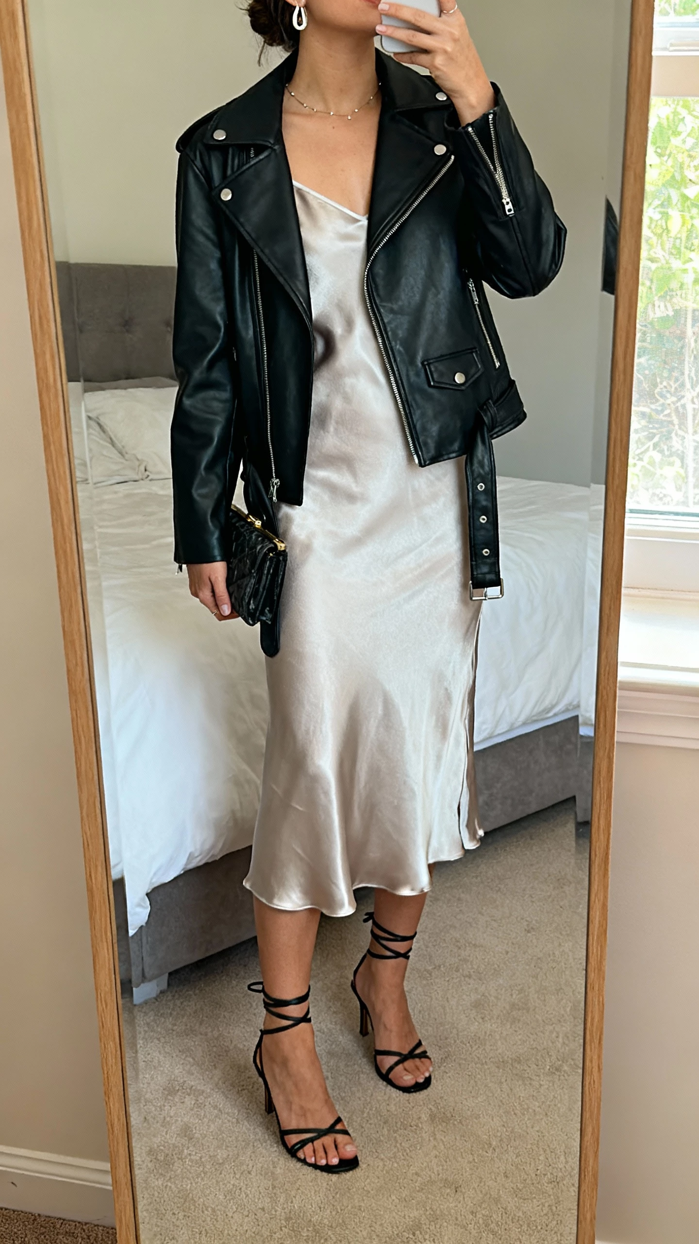 iPhone mirror selfie of a woman wearing a black leather jacket over a silky slip dress with strappy heels, delicate earrings, and a small clutch, face not visible, cropped at neck, bedroom full-length mirror, soft window light, natural iPhone photo quality