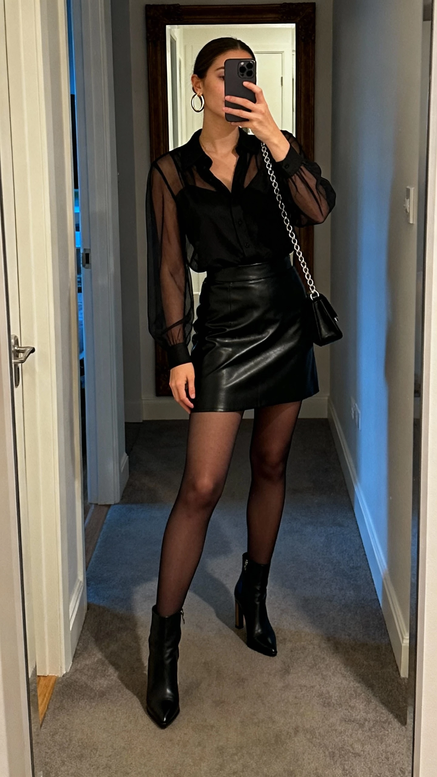 iPhone mirror selfie of a woman wearing a black leather skirt with a sheer-sleeve blouse, opaque tights, heeled ankle boots, hoop earrings, and a chain-strap bag, face not visible, full-length mirror in entryway, evening indoor lighting, natural iPhone photo quality.