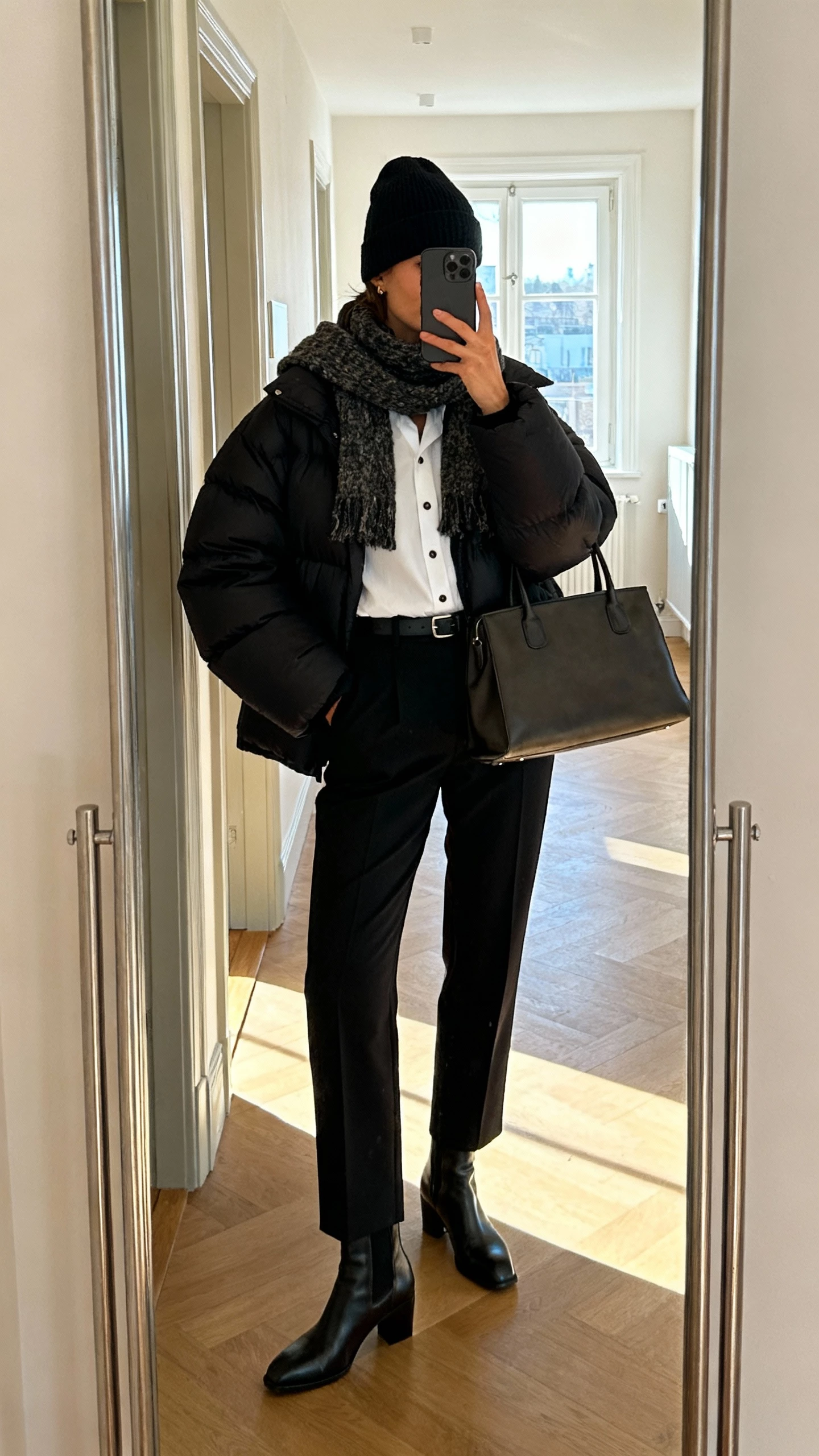 iPhone mirror selfie of a woman wearing a black oversized puffer jacket over a crisp white button-down tucked into tailored black trousers, chunky scarf, beanie, sleek ankle boots, structured tote, face not visible, cropped at neck, hallway mirror in an apartment, natural morning window light, natural iPhone photo quality