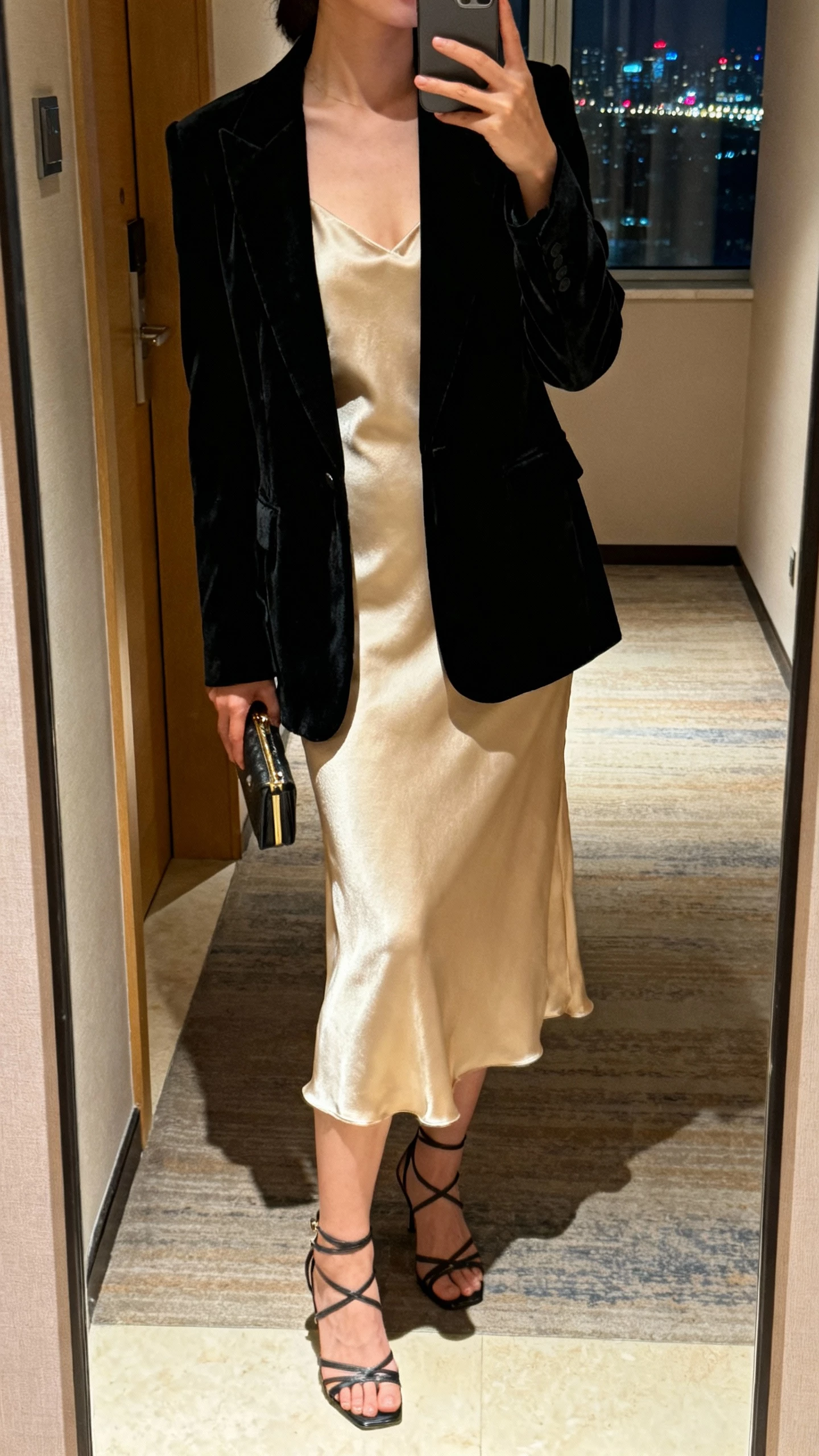 iPhone mirror selfie of a woman wearing a black velvet blazer over a champagne slip dress with strappy heels and a small clutch, face not visible, cropped at neck, hotel hallway mirror at night with city lights through a window, natural iPhone photo quality