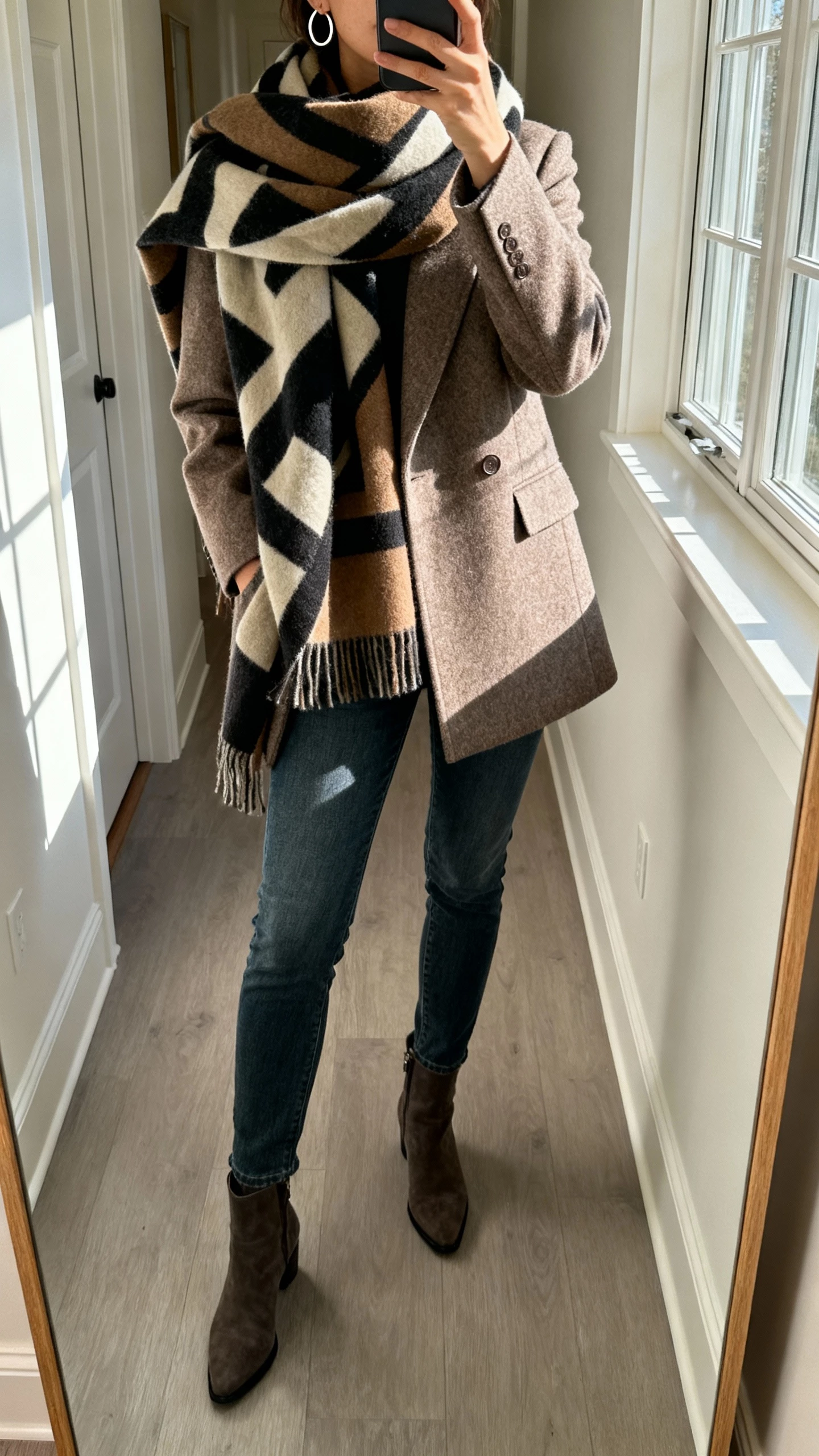 iPhone mirror selfie of a woman wearing a bold oversized statement scarf wrapped over a tailored wool coat, slim jeans, ankle boots, simple hoop earrings, face not visible, cropped at neck, hallway mirror with morning window light, natural iPhone photo quality.