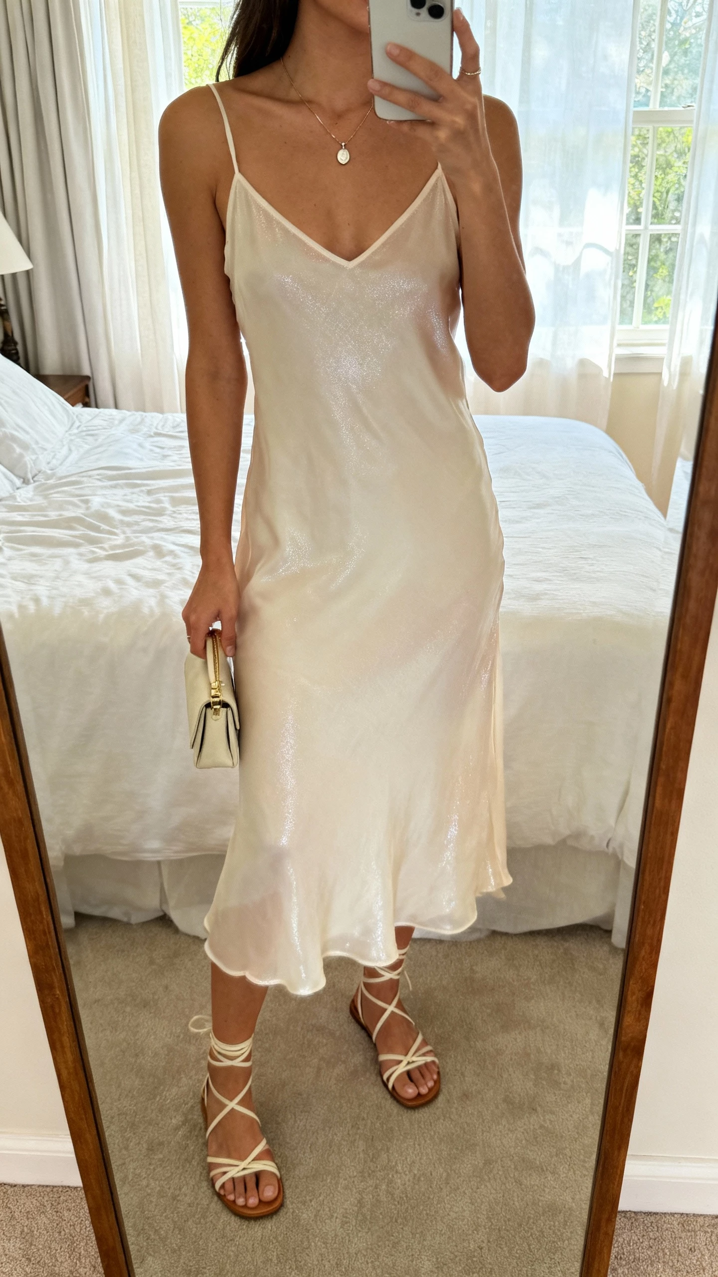 iPhone mirror selfie of a woman wearing a breezy slip dress with subtle, barely-there shine, strappy sandals, delicate pendant necklace, small clutch, face not visible, cropped at neck, bedroom mirror, soft window light, natural iPhone photo quality.