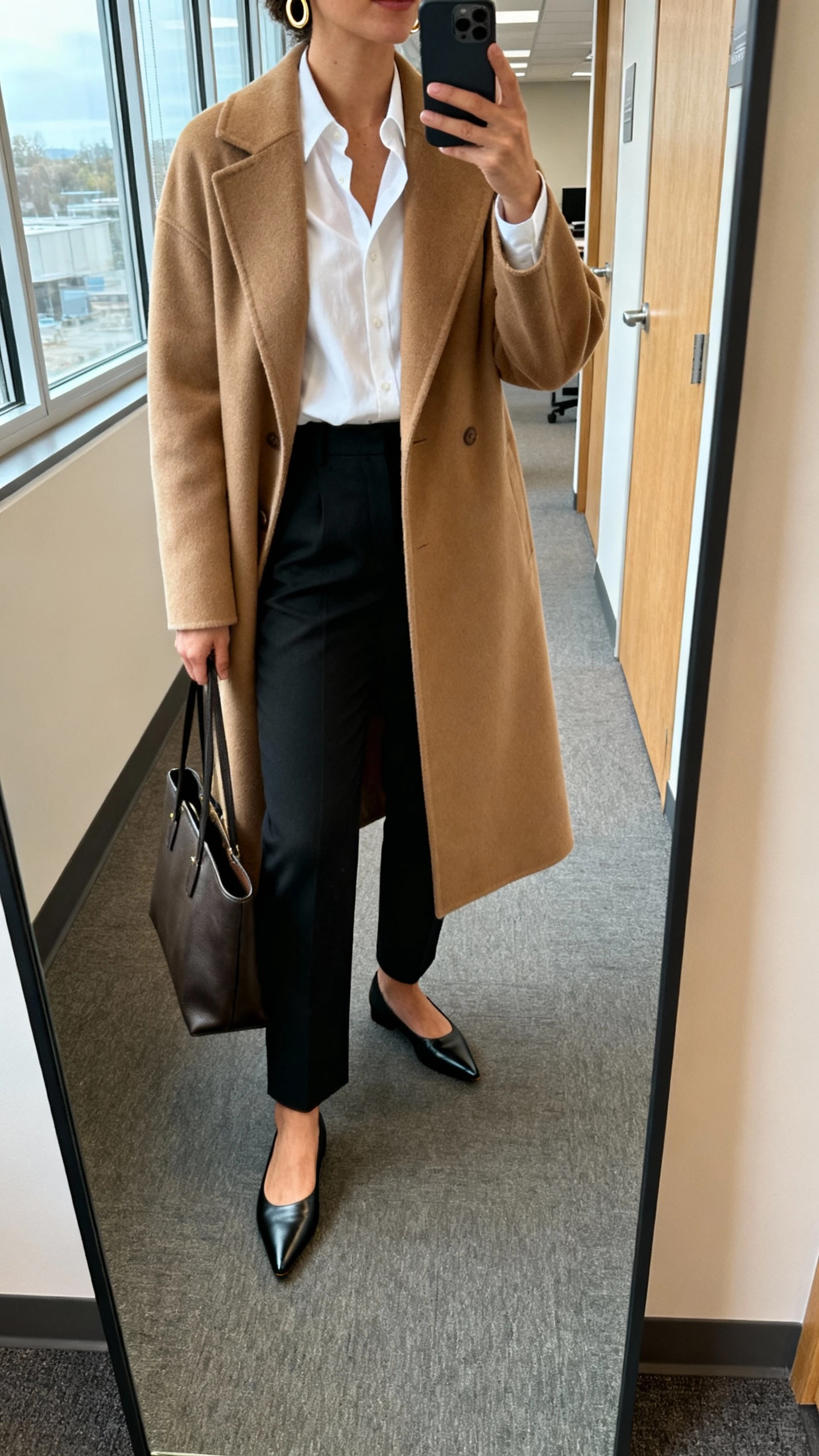 iPhone mirror selfie of a woman wearing a camel coat over a crisp white blouse, tailored black trousers, pointed loafers, structured tote, simple gold hoops, face not visible, cropped at neck, office hallway mirror, morning window light, natural iPhone photo quality.