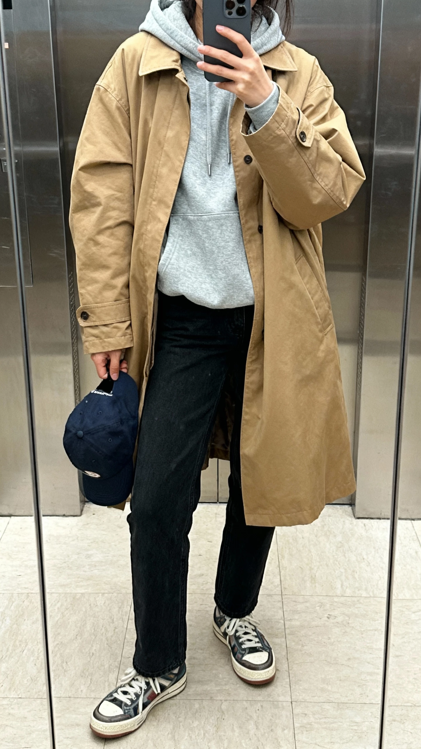 iPhone mirror selfie of a woman wearing a camel dad coat layered over a grey hoodie, black straight jeans, retro sneakers, baseball cap in hand, face not visible, elevator mirror, afternoon light, casual iPhone photo