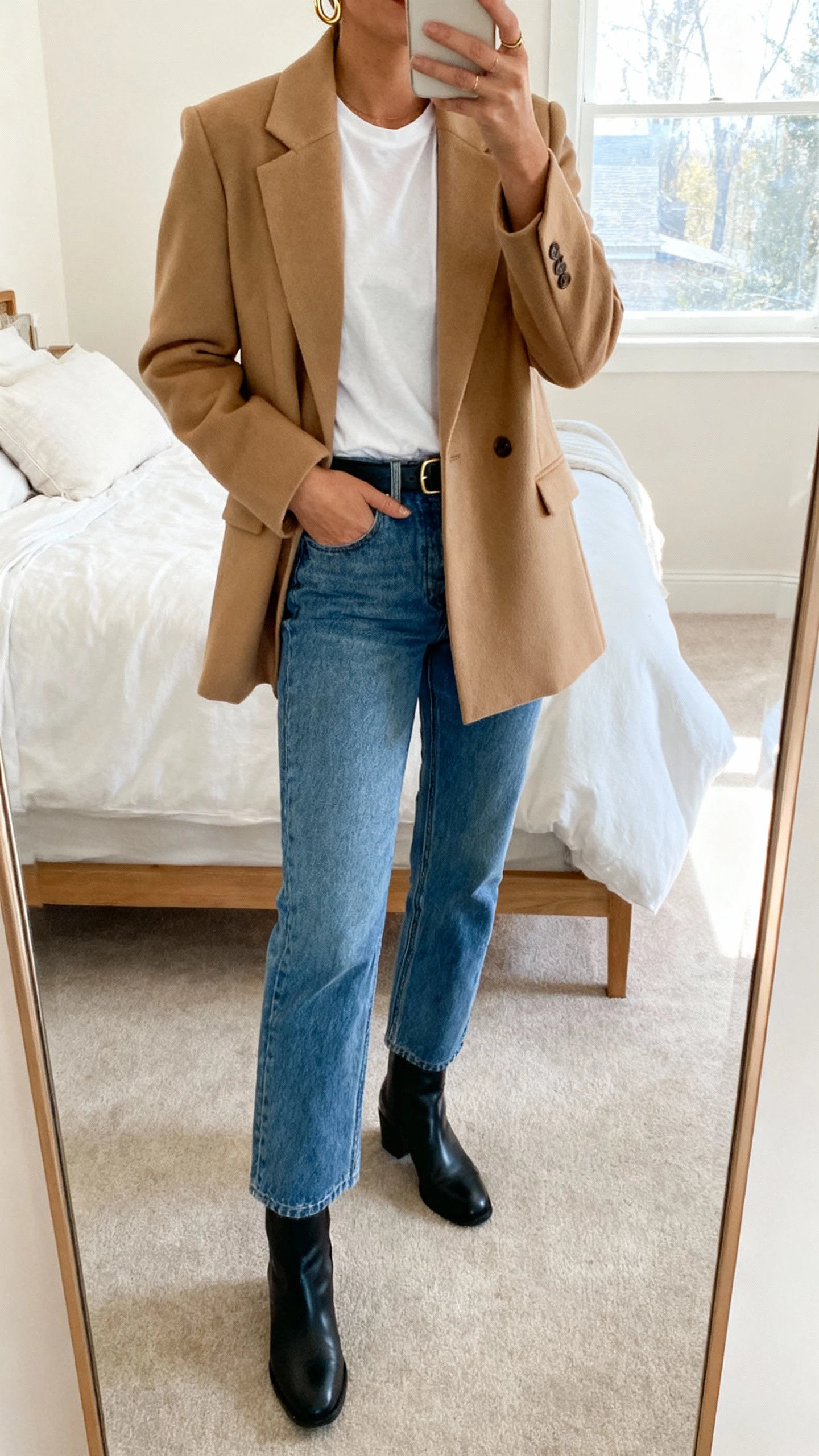 iPhone mirror selfie of a woman wearing a camel tailored coat over a white tee, straight blue denim, black leather ankle boots, simple belt, minimal gold hoops, face not visible, cropped at neck, bedroom mirror with morning window light, natural iPhone photo quality.