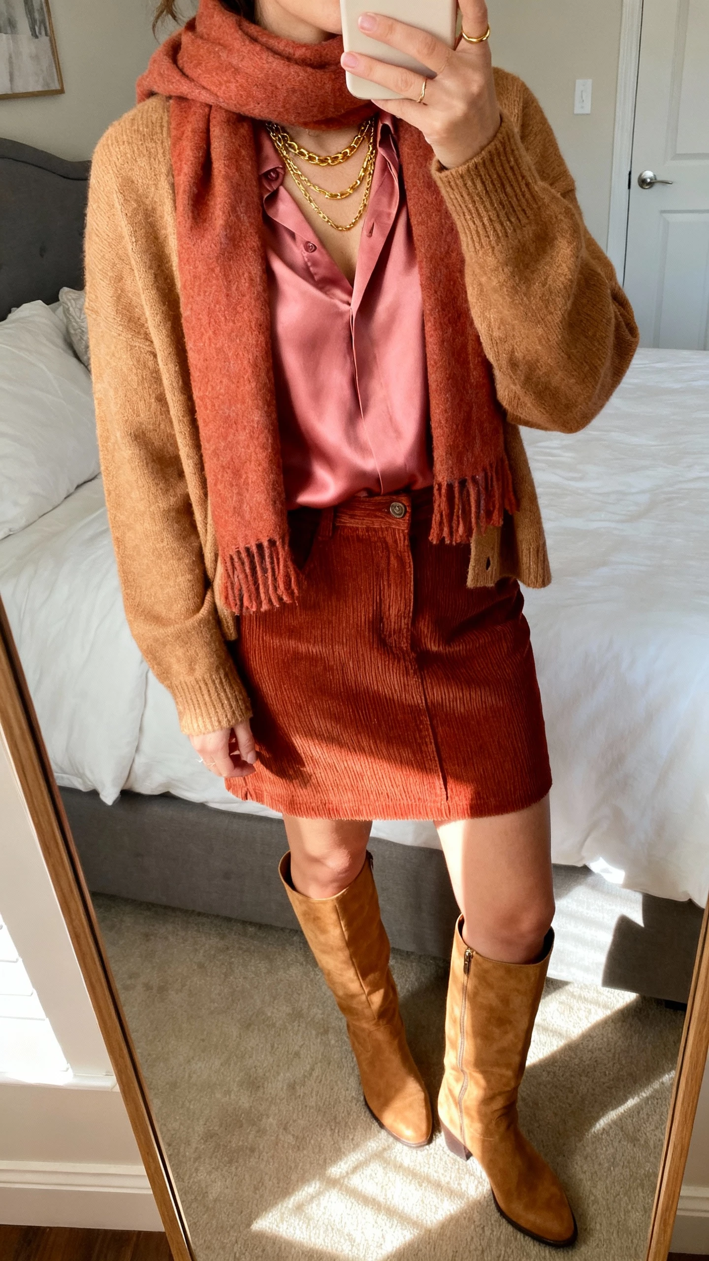 iPhone mirror selfie of a woman wearing a caramel cardigan over a rose silk blouse, rust corduroy skirt, tan knee-high boots, warm-toned scarf, gold layered necklaces, face not visible, bedroom mirror with warm afternoon light, casual iPhone quality