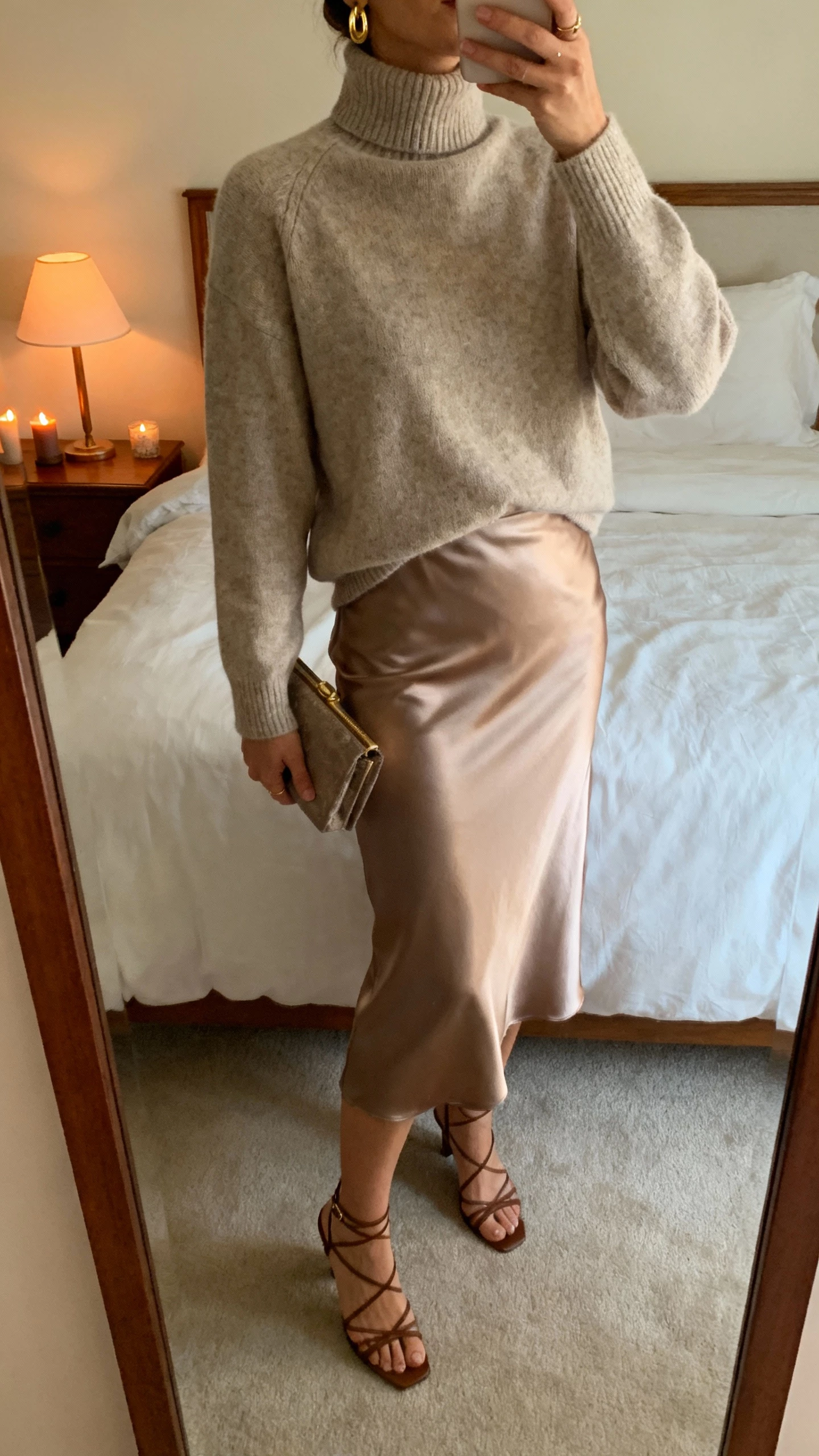 iPhone mirror selfie of a woman wearing a cashmere turtleneck, satin midi skirt, strappy heels, delicate gold earrings, small clutch, face not visible, cropped at neck, bedroom mirror with warm lamp light that feels like candlelight, natural iPhone photo quality
