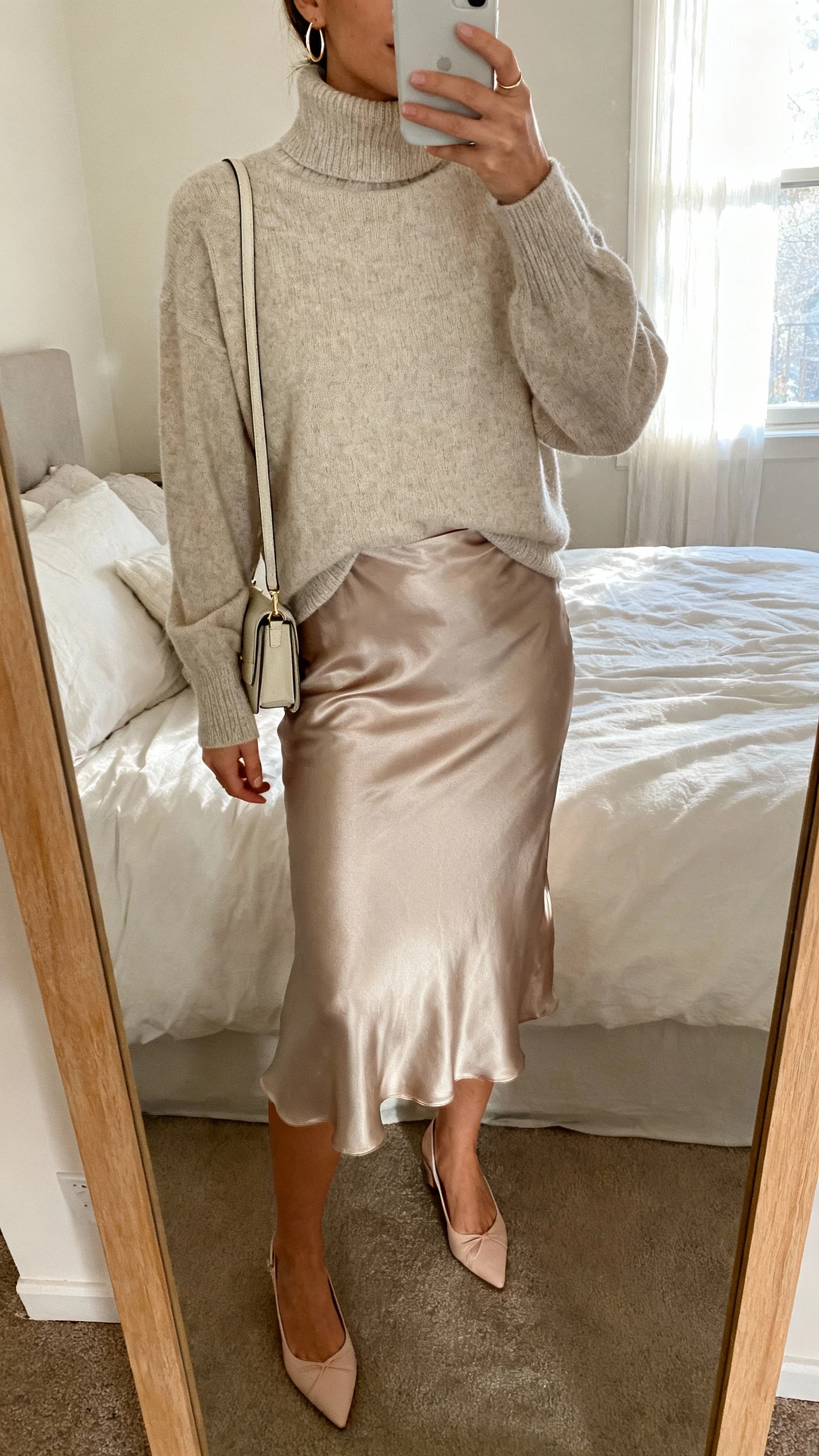 iPhone mirror selfie of a woman wearing a cashmere turtleneck tucked into a satin slip skirt with delicate heels, small shoulder bag, dainty hoop earrings, face not visible, cropped at neck, bedroom mirror with window light, natural iPhone photo quality.