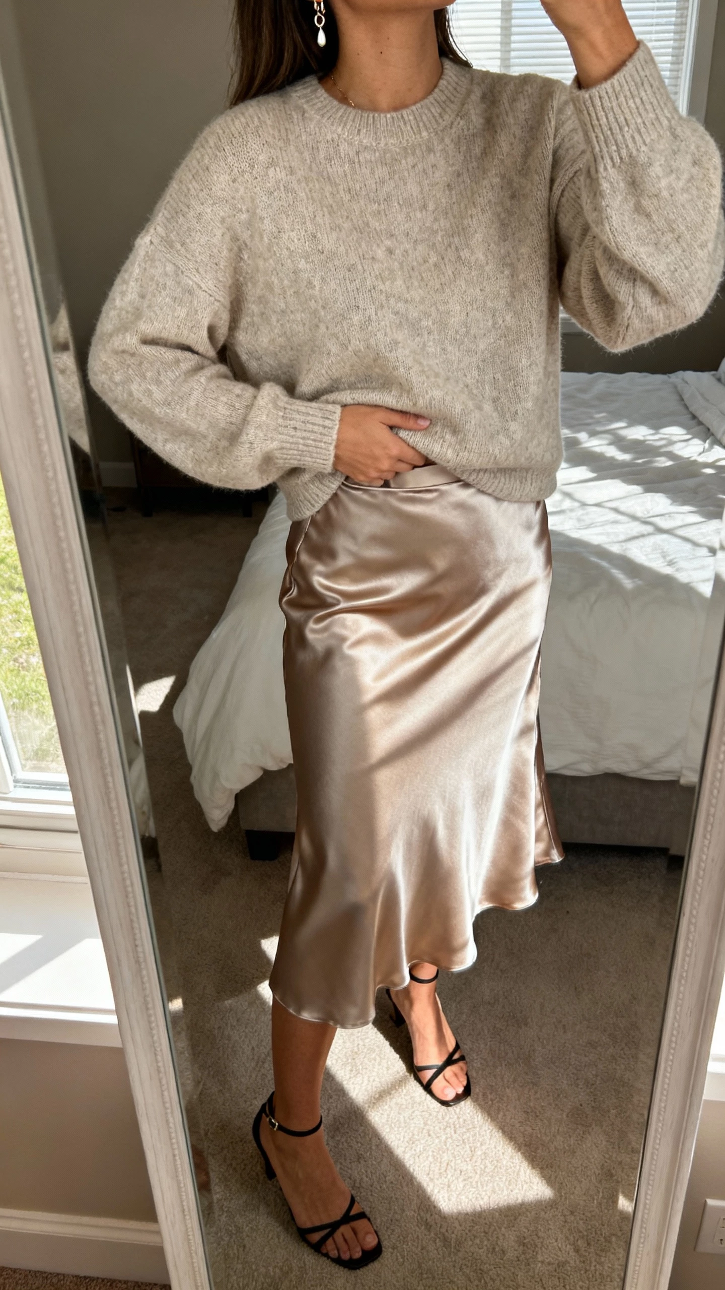 iPhone mirror selfie of a woman wearing a cashmere sweater tucked into a satin midi skirt with delicate earrings and ankle-strap heels, face not visible, cropped at neck, bedroom mirror with morning window light, natural iPhone photo quality.