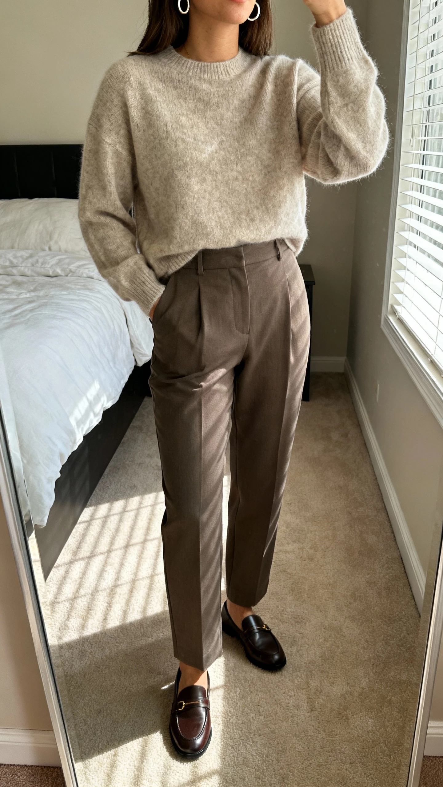 iPhone mirror selfie of a woman wearing a cashmere-look sweater tucked into tailored trousers with simple hoop earrings and loafers, face not visible, cropped at neck, bedroom mirror with morning window light, natural iPhone photo quality.