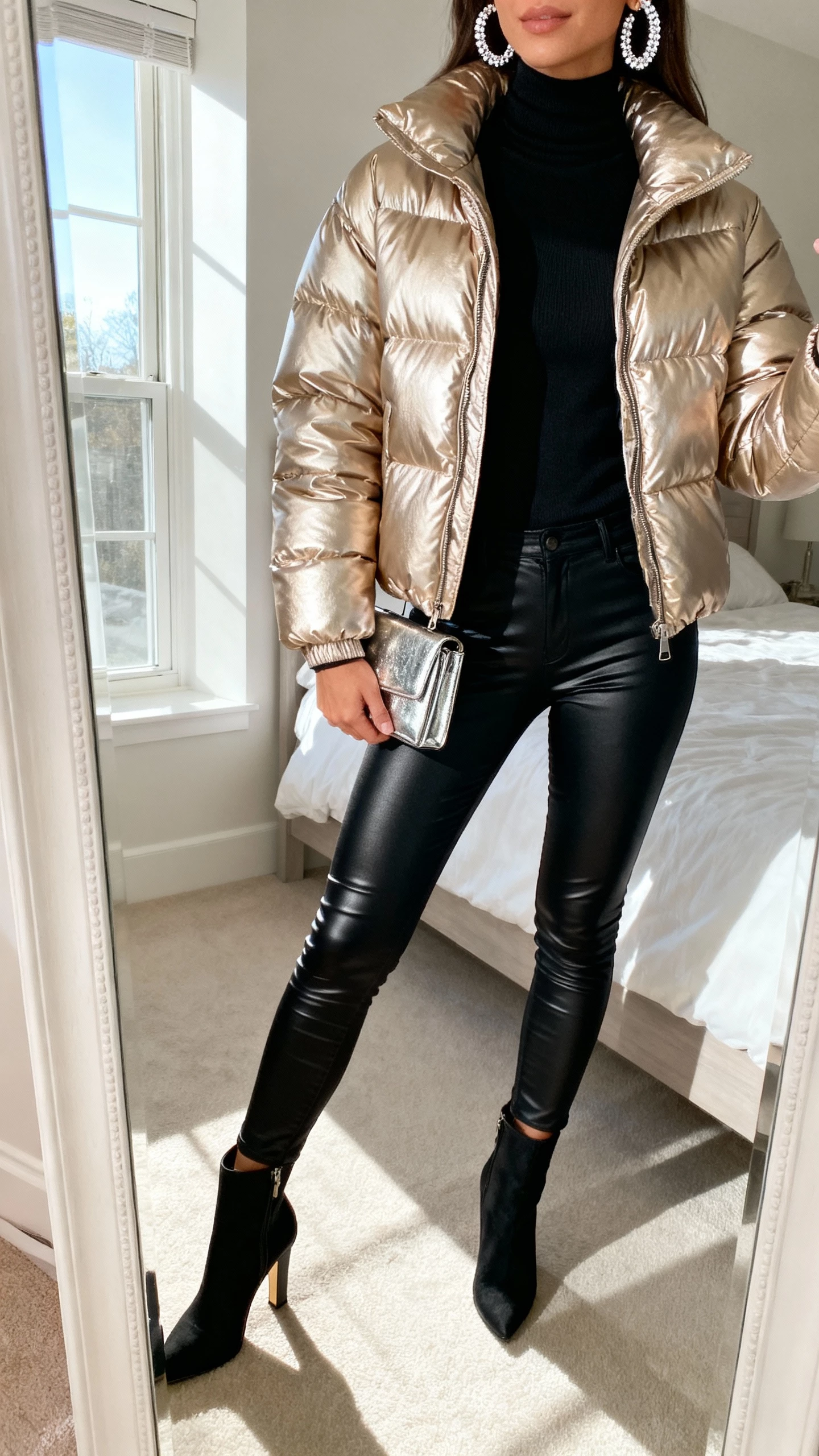 iPhone mirror selfie of a woman wearing a champagne metallic puffer jacket over a slim black turtleneck, black coated skinny jeans, heeled ankle boots, sparkly hoop earrings, a small metallic clutch, face not visible, cropped at neck, bedroom mirror with natural window light, natural iPhone photo quality
