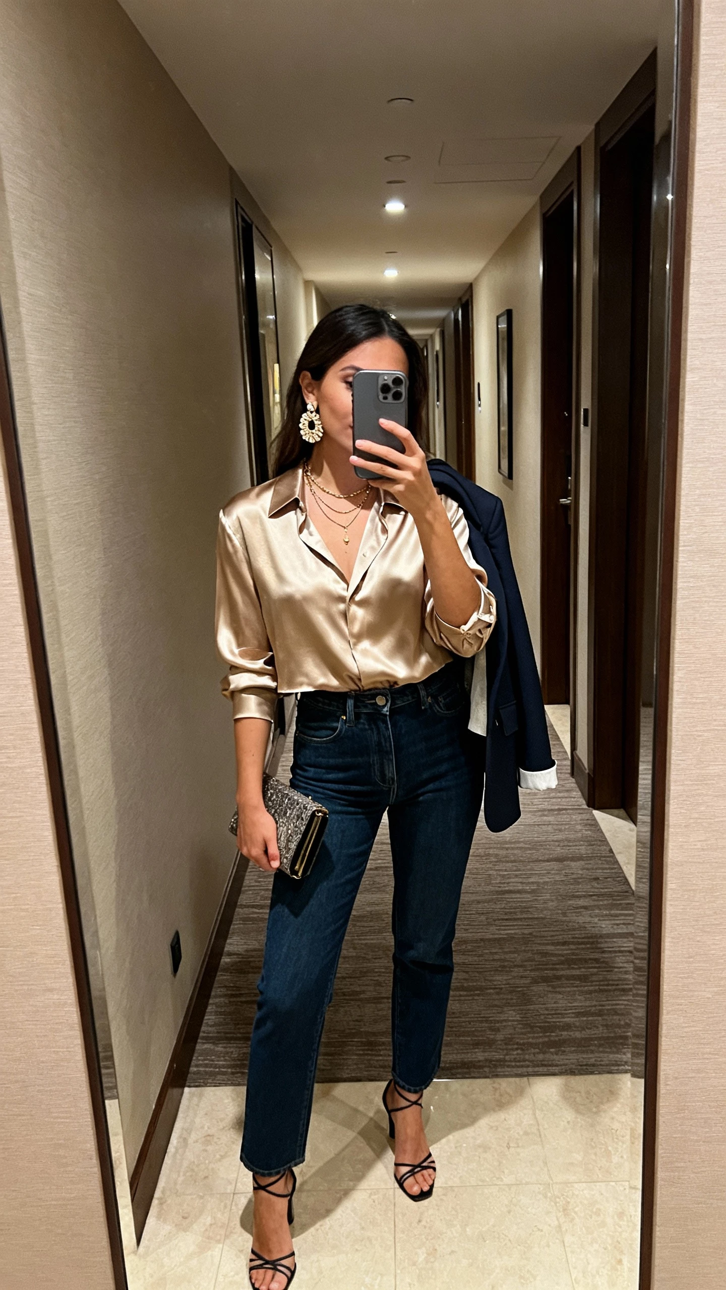 iPhone mirror selfie of a woman wearing a champagne silk blouse tucked into dark blue high-waisted straight jeans, strappy heels, delicate layered necklaces, statement earrings, and a clutch, with a cropped blazer over shoulder; hotel hallway mirror, evening indoor light, face not visible, natural iPhone aesthetic.