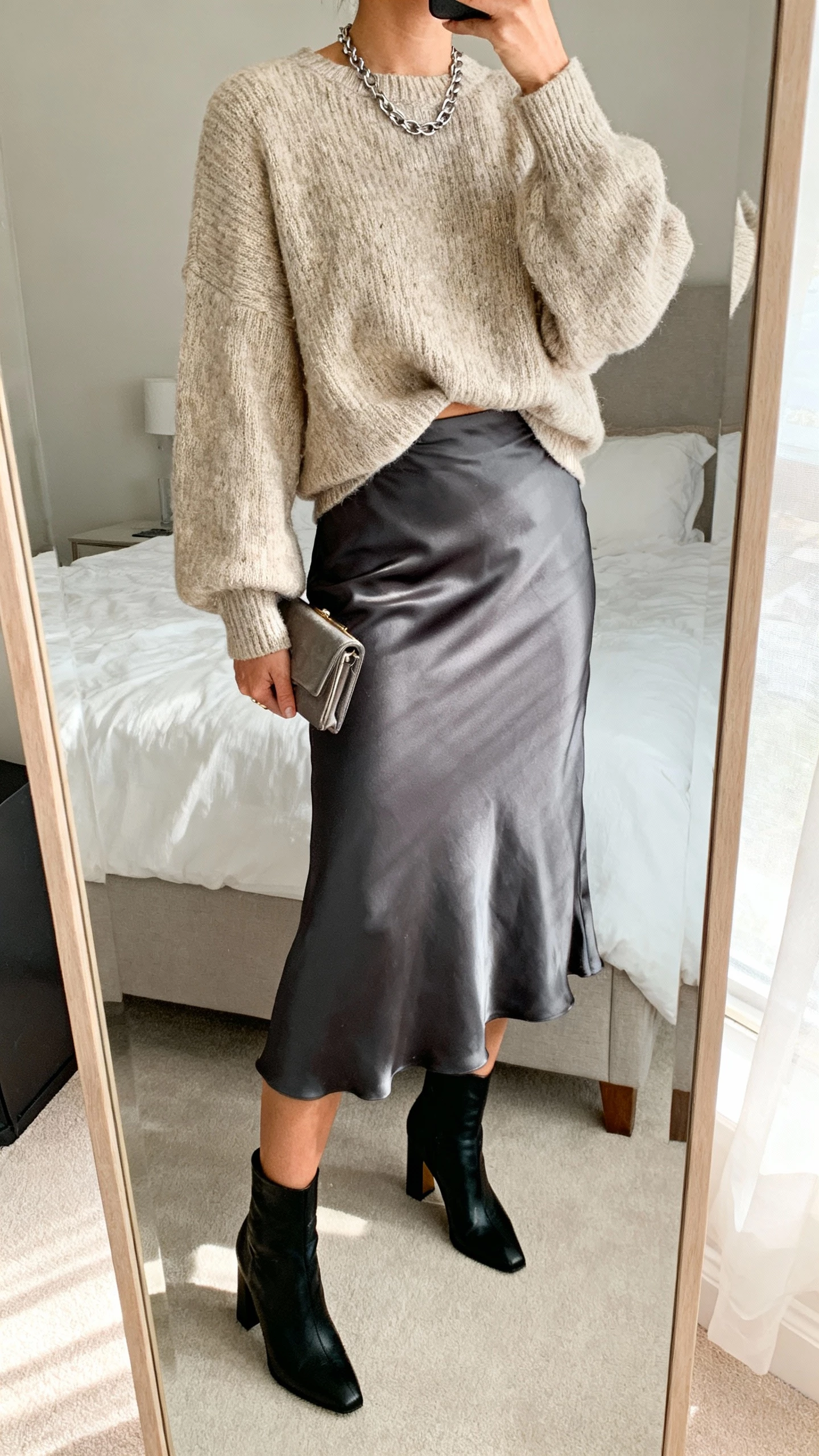 iPhone mirror selfie of a woman wearing a chunky oatmeal sweater half-tucked into a charcoal satin midi skirt, black heeled ankle boots, simple chain necklace, small clutch, face not visible, bedroom full-length mirror with soft morning light, natural iPhone photo quality.