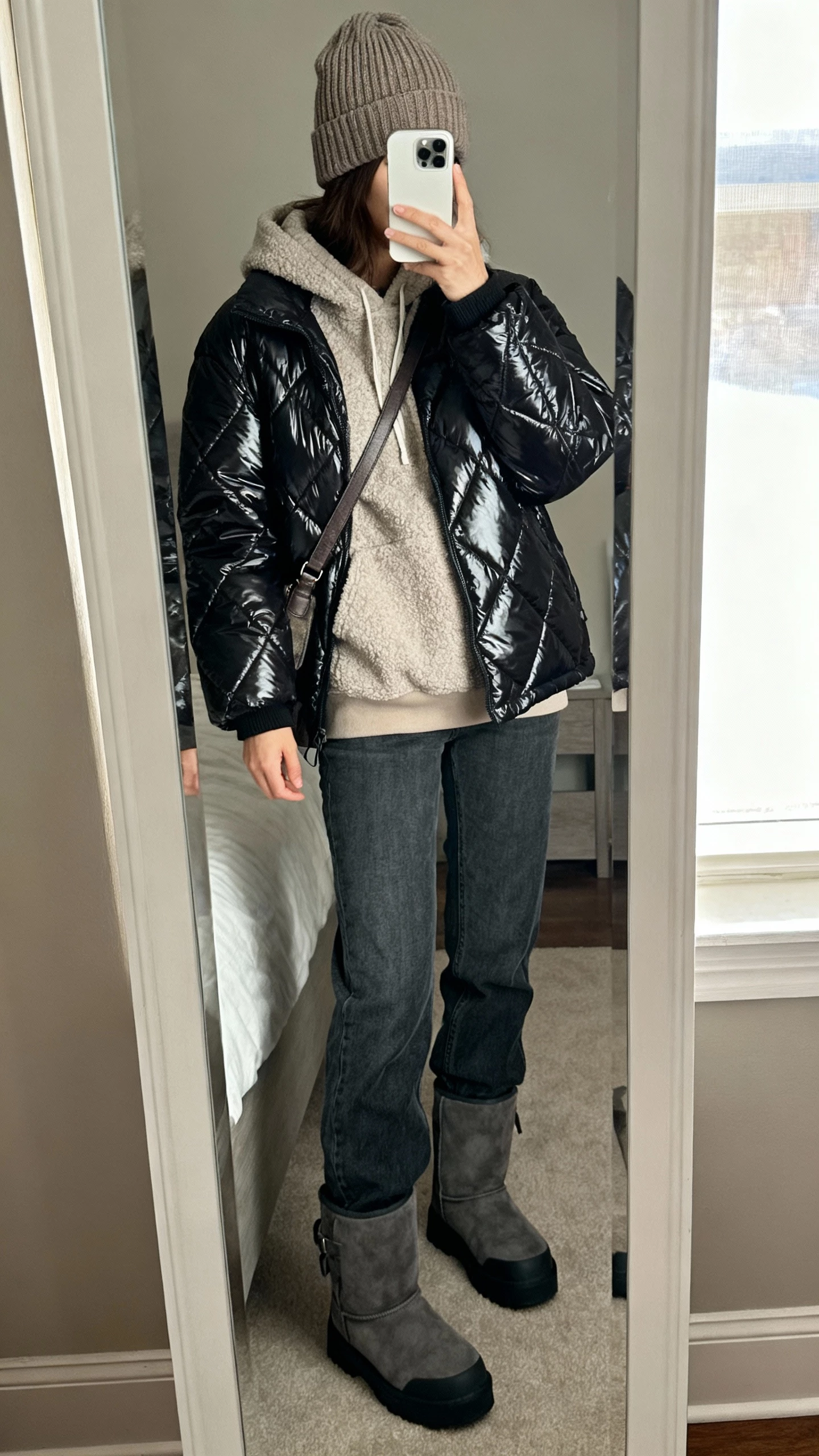 iPhone mirror selfie of a woman wearing a chunky ribbed beanie, glossy quilted puffer jacket, fleece hoodie, straight-leg jeans, chunky winter boots, and a crossbody bag; face not visible, cropped at neck, bedroom mirror with window light, natural iPhone photo quality.