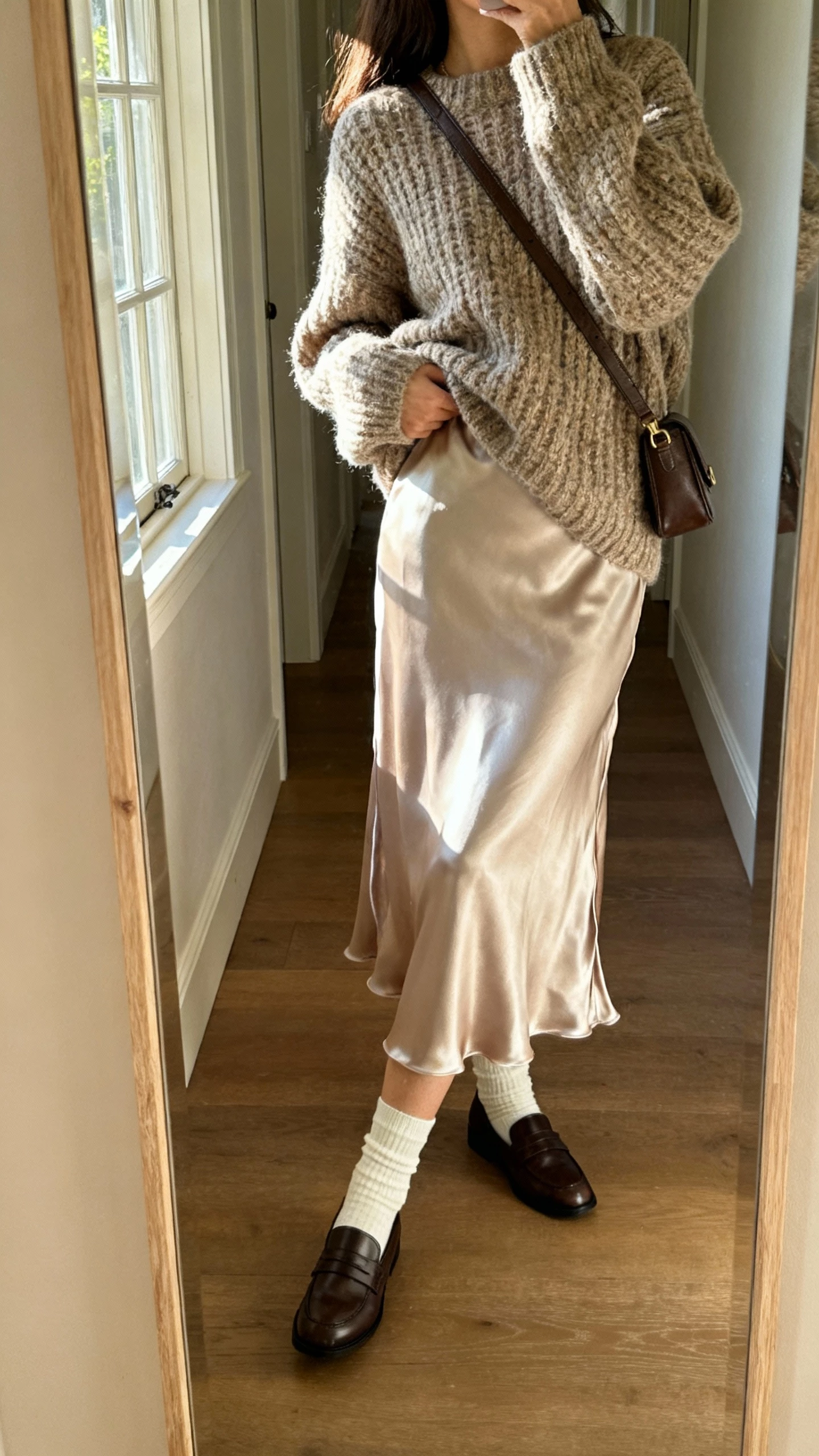 iPhone mirror selfie of a woman wearing a chunky sweater layered over a satin slip dress, with socks and loafers and a crossbody bag, face not visible, side angle in hallway mirror, soft afternoon window light, natural iPhone photo.