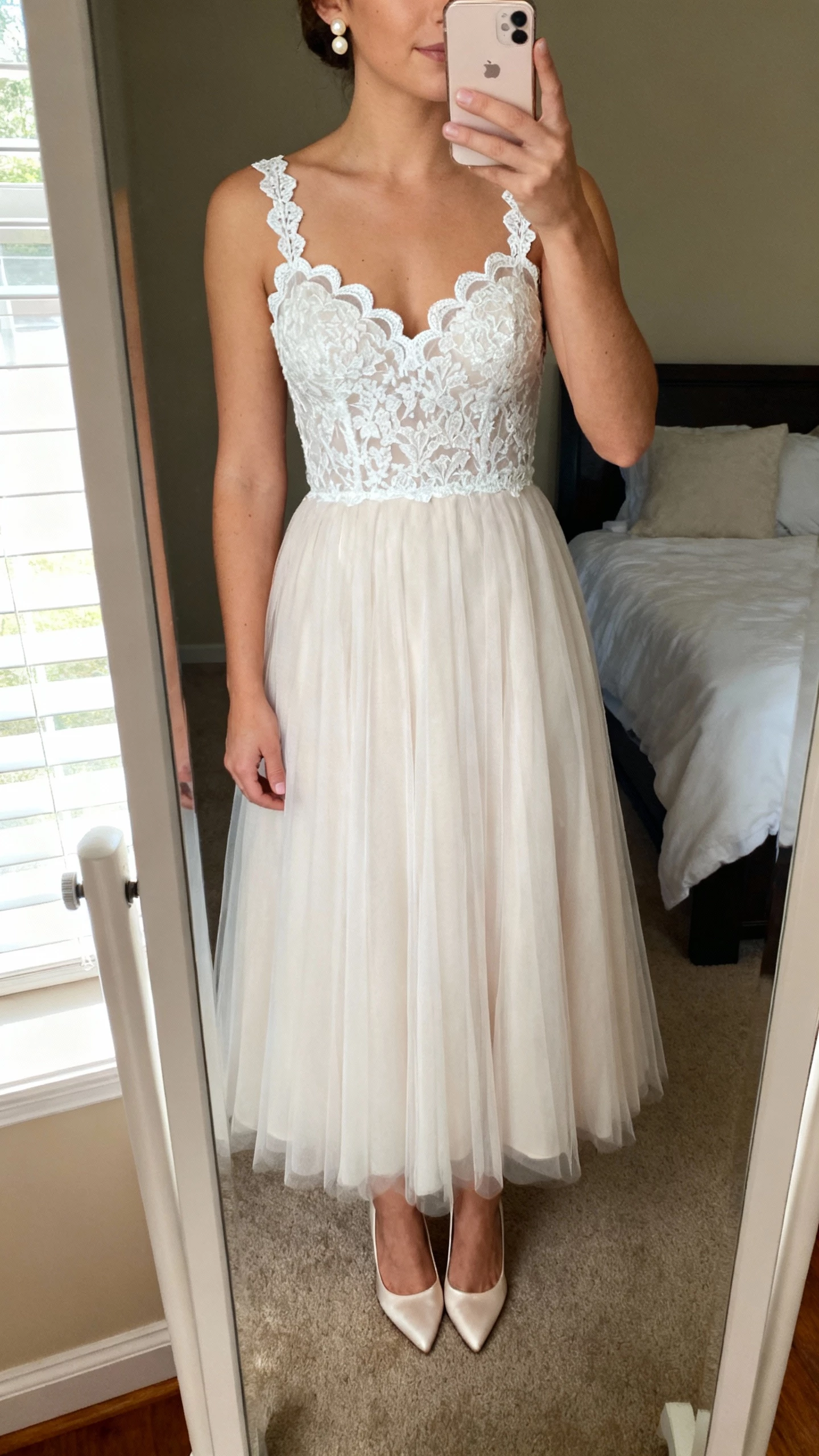 iPhone mirror selfie of a woman wearing a classic lace A-line wedding dress with scalloped lace bodice, illusion lace straps, soft tulle skirt, pearl stud earrings, and satin pumps; face not visible, cropped at neck, bedroom mirror with morning window light, natural iPhone photo quality.