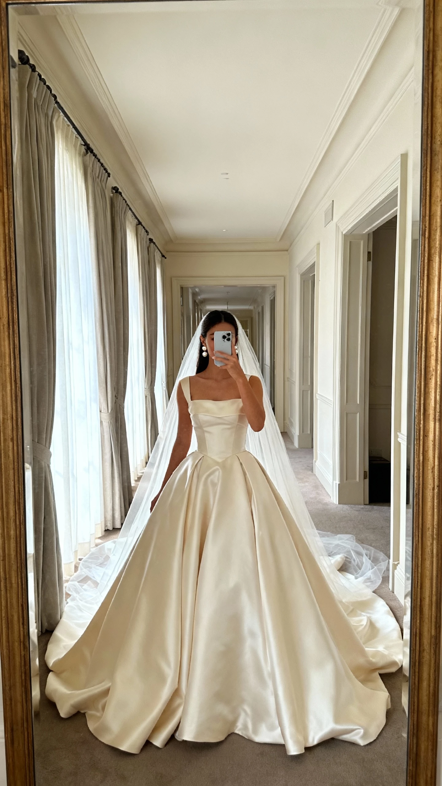 iPhone mirror selfie of a woman wearing a classic cathedral ball gown with structured satin bodice, full voluminous skirt, long cathedral train, pearl earrings, and long veil, face not visible, cropped at neck, grand hallway mirror, soft window light, natural iPhone photo quality