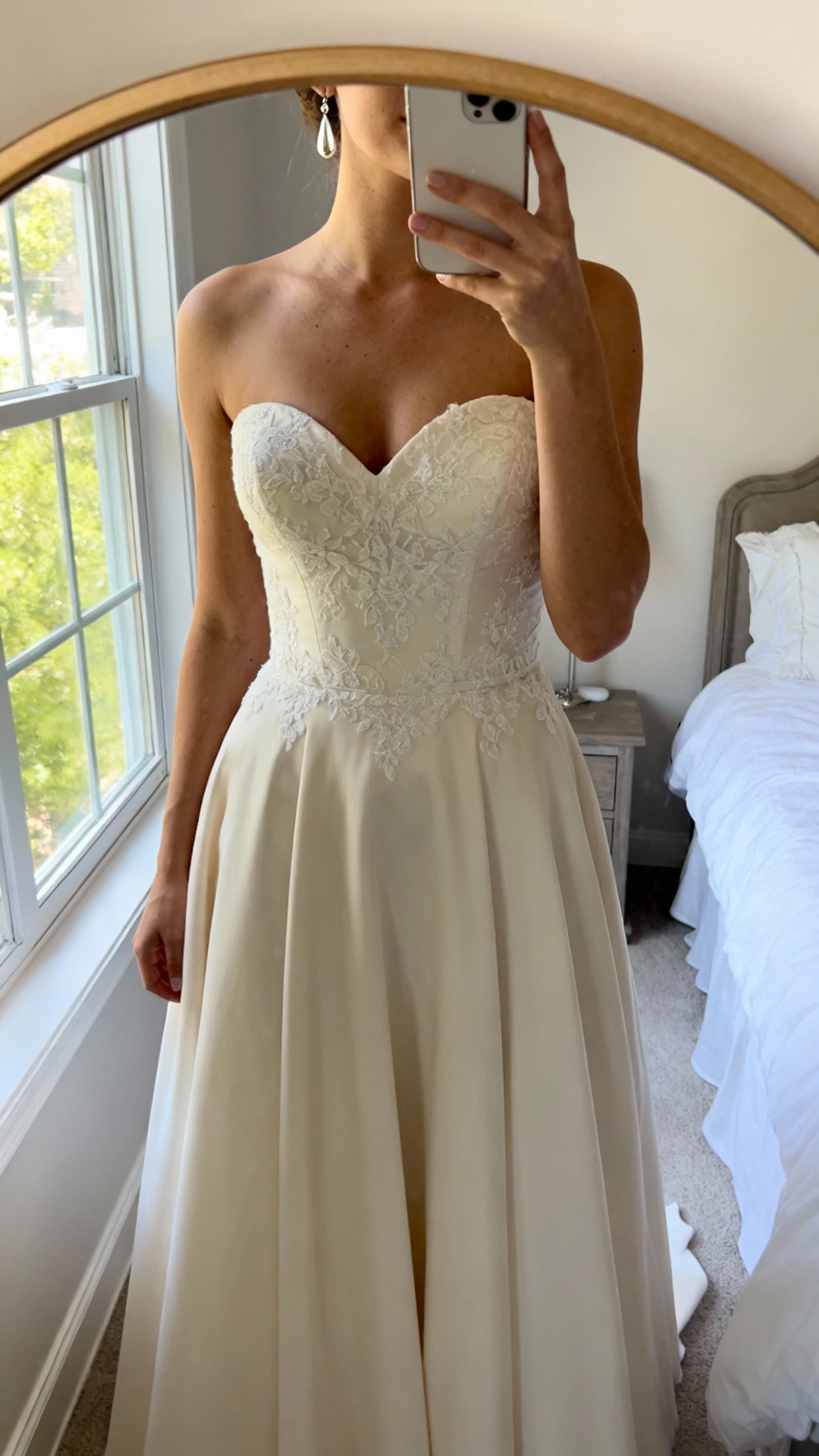 iPhone mirror selfie of a woman wearing a classic sweetheart-neckline wedding dress with a fitted bodice, soft A-line skirt, delicate lace detailing, simple drop earrings, face not visible, bedroom mirror, natural window light, natural iPhone photo quality.