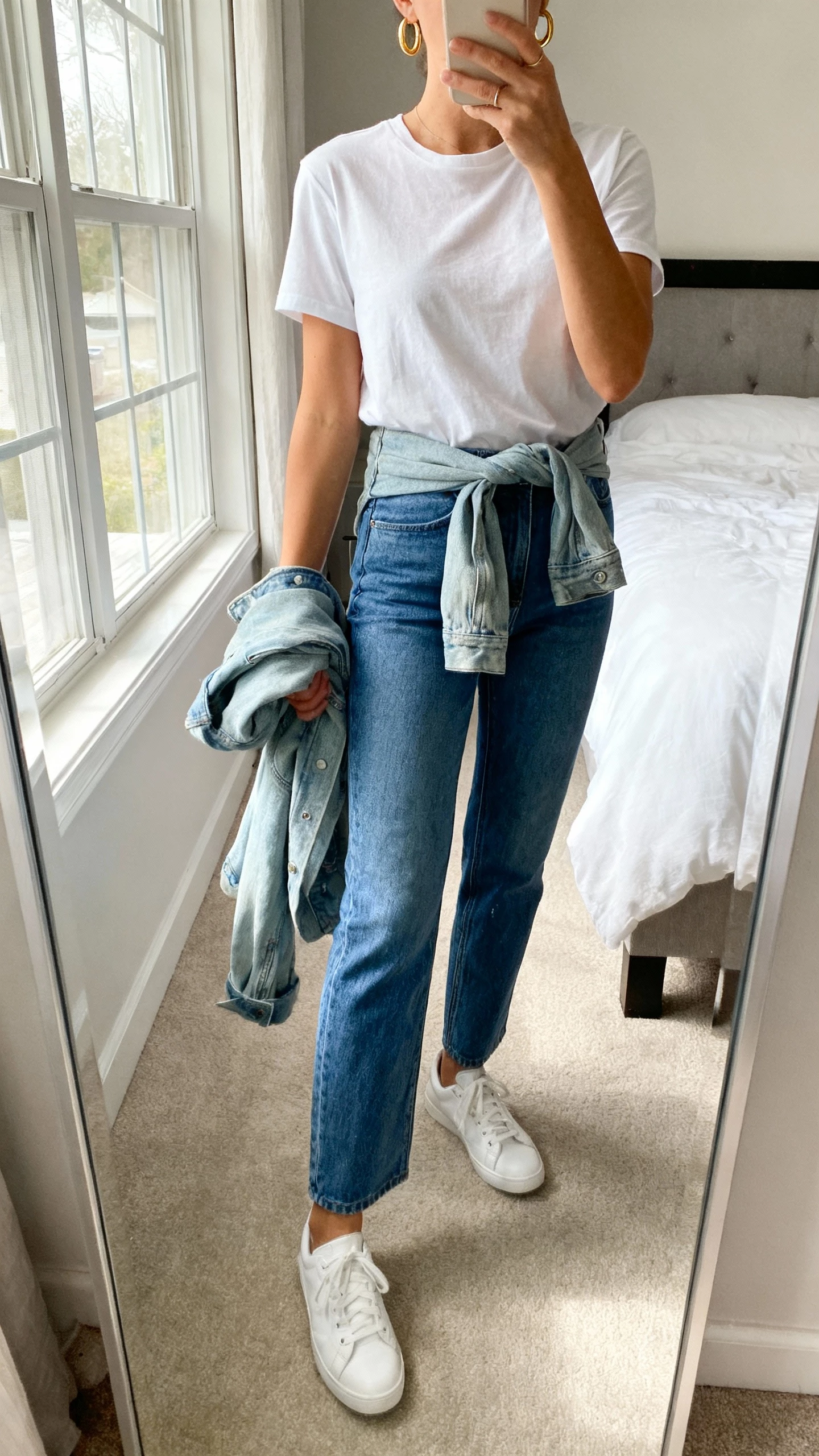 iPhone mirror selfie of a woman wearing a classic white tee tucked into straight-leg blue jeans, white sneakers, simple gold hoops, light-wash denim jacket tied at the waist or draped over arm, face not visible, cropped at neck, bedroom mirror with morning window light, natural iPhone photo quality.