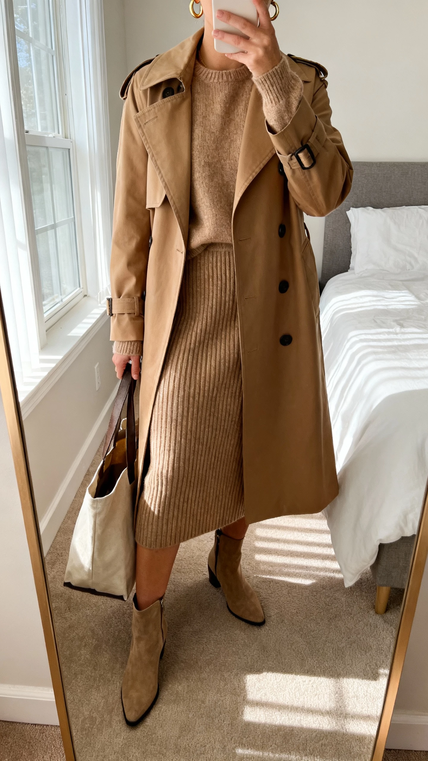 iPhone mirror selfie of a woman wearing a classic camel trench coat over a matching ribbed knit set (sweater and skirt), ankle boots, simple gold hoops, tote bag in hand, face not visible, cropped at neck, bedroom mirror with morning window light, natural iPhone photo quality